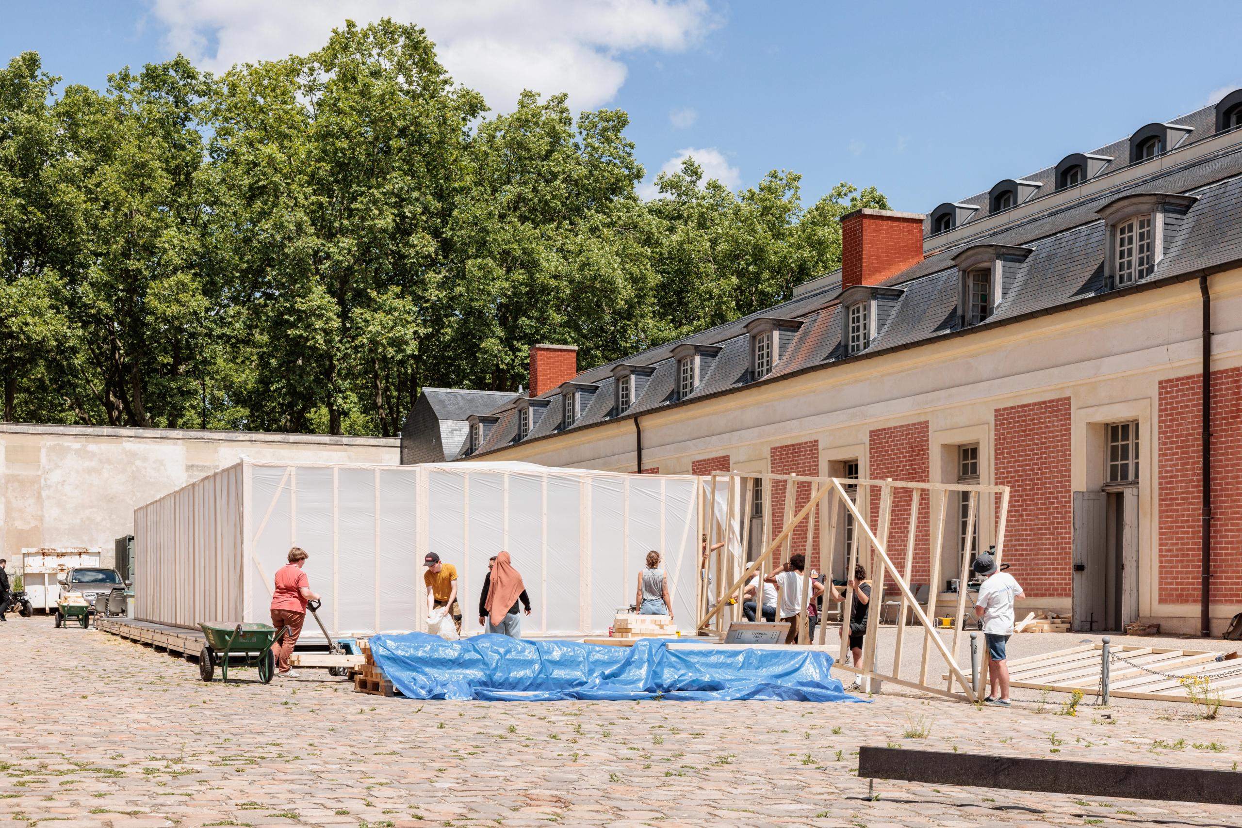 Workshop 60x60 à Versailles : montage d’une ossature bois modulaire en panneaux manuportables, fermés par film plastique translucide, dans la cour pavée de la Maréchalerie.