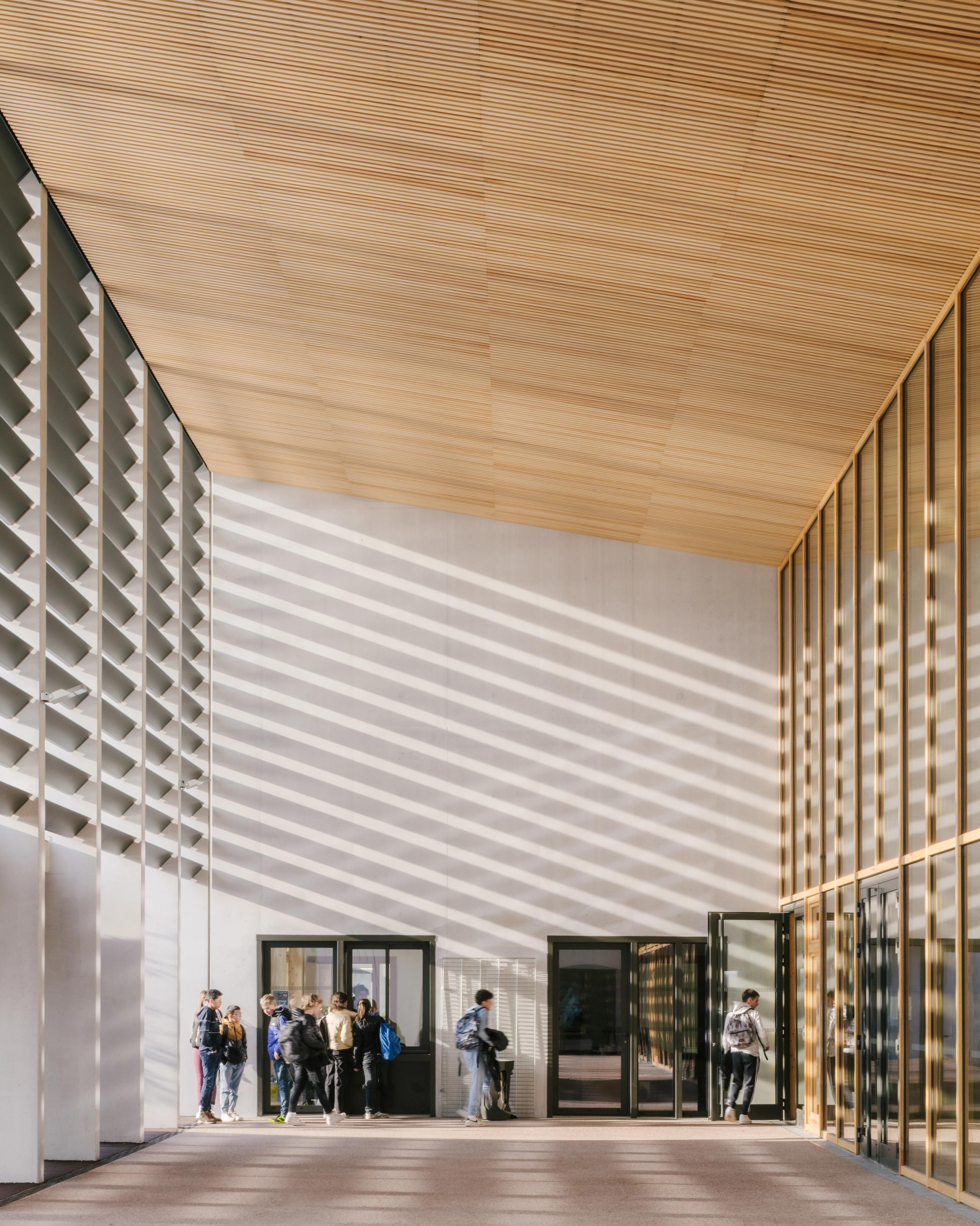 Portique d’entrée du Collège du Ban à Vagney, vaste préau bois-béton baigné de lumière, rythmé par brise-soleil verticaux et façade vitrée sur la cour.