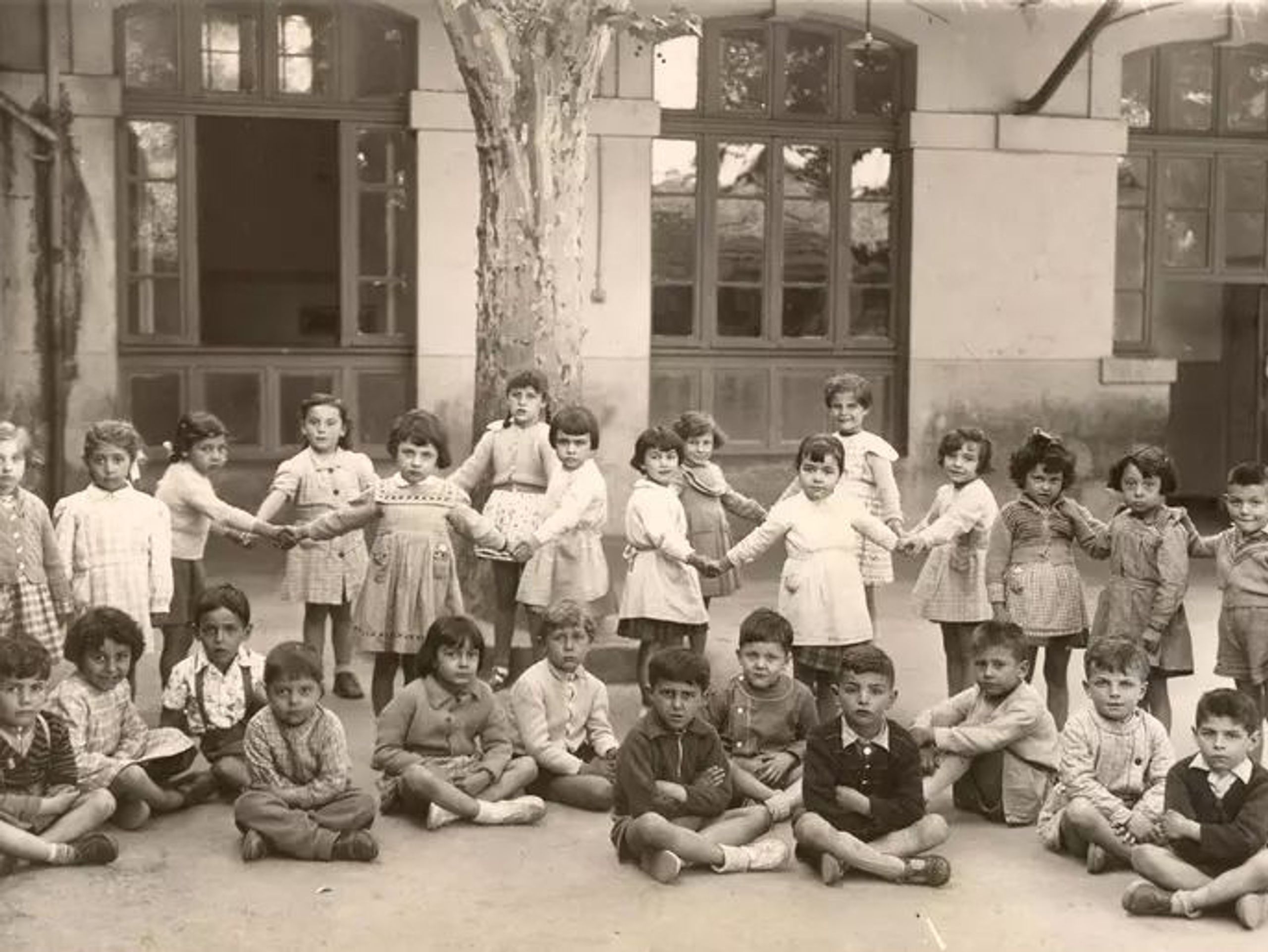 Photographie d’archive d’une cour d’école à Marseille : façade enduite à grandes baies vitrées bois, soubassement sombre et arbre central structurant l’espace.