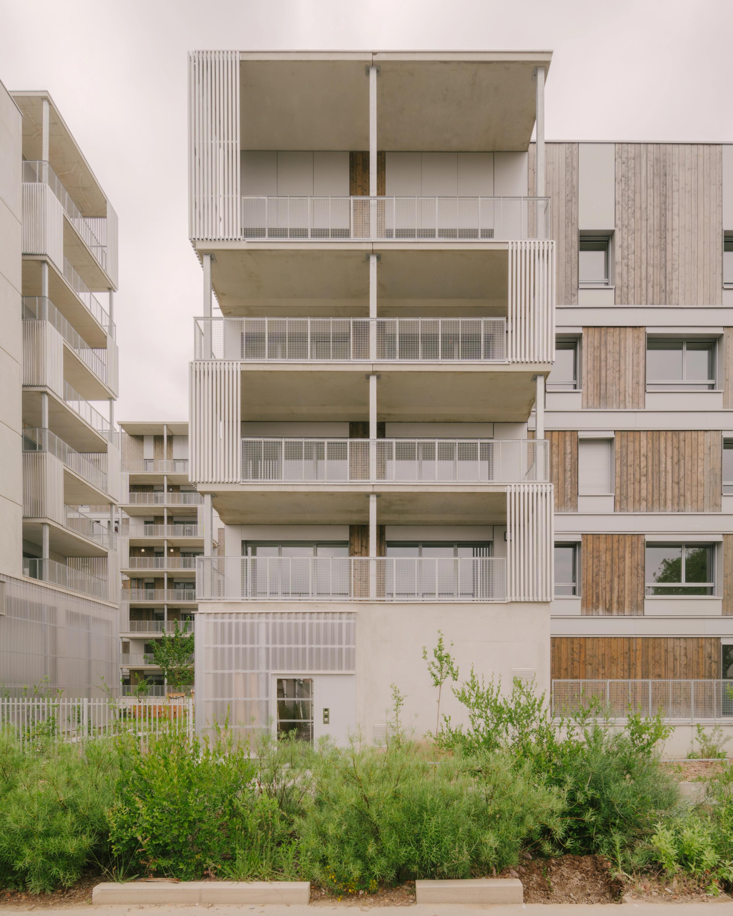 Vue de la façade des 116 logements à Vénissieux par Tectoniques, volumes fragmentés, balcons filants, ossature bois-béton et exosquelettes métalliques sur jardin planté.