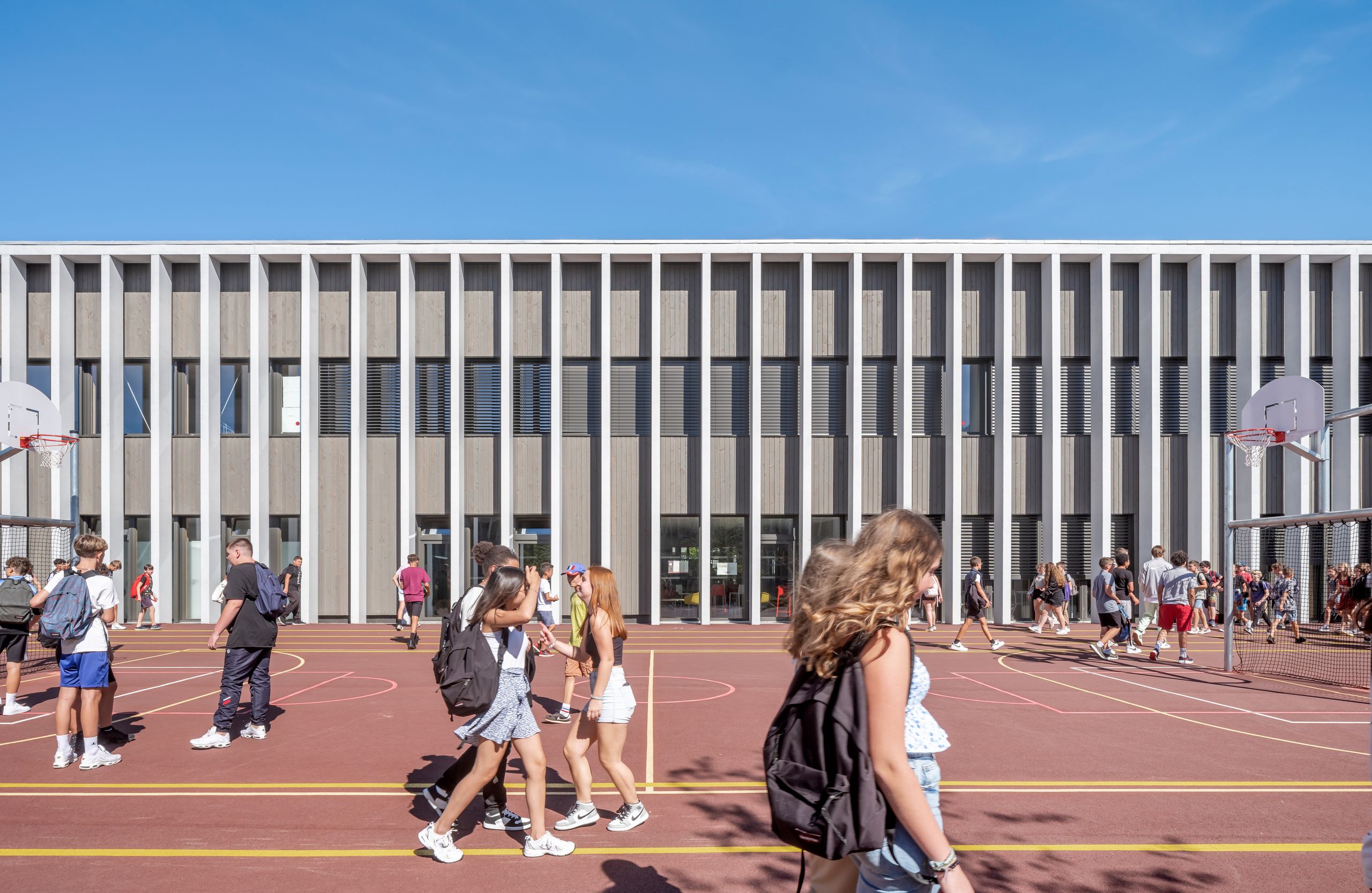 Façade rythmée en bois et béton du Collège du Ban à Vagney, cadrant la cour rouge du plateau sportif animé par les élèves en extérieur.