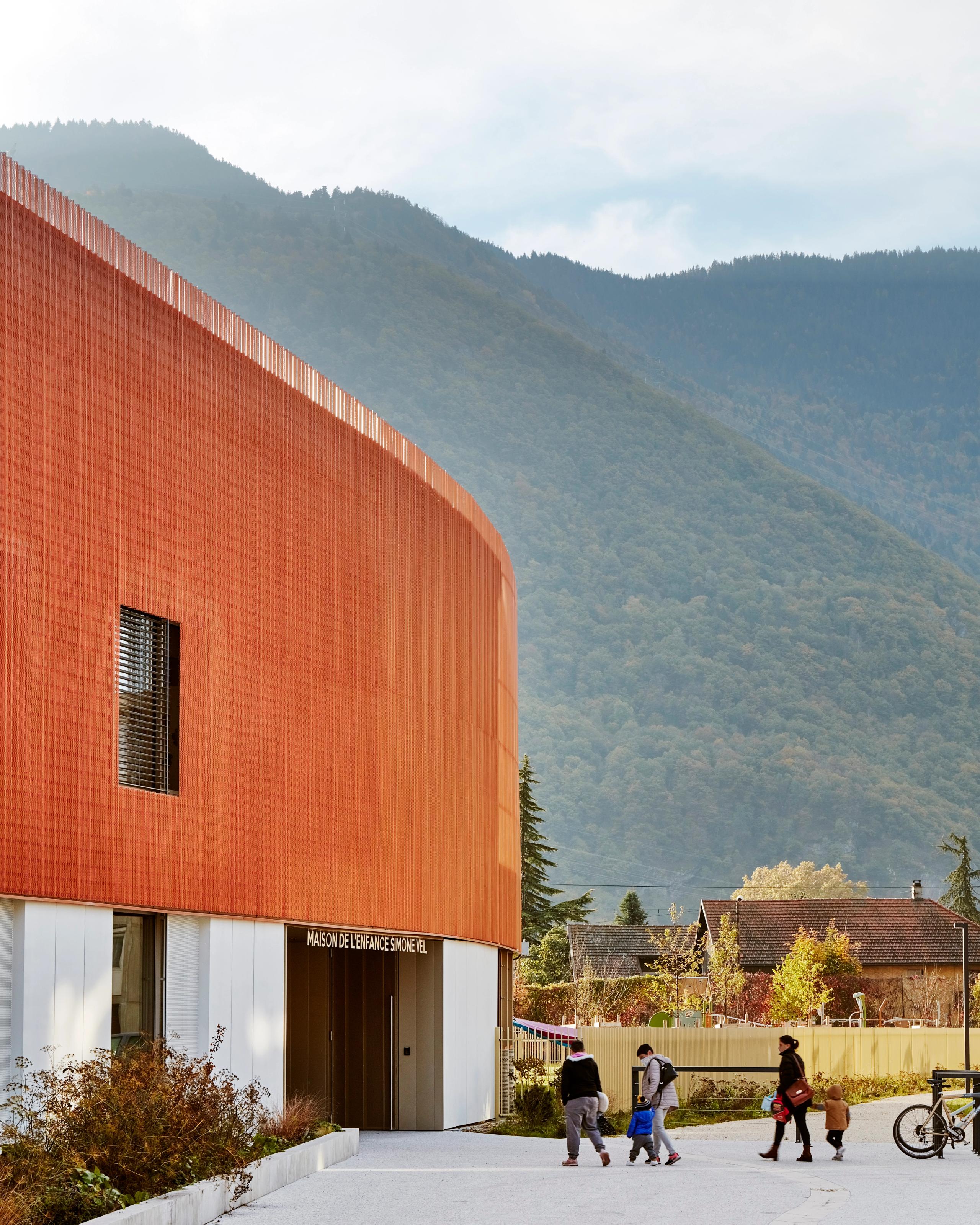 Façade courbe en métal cuivré de la Maison de l’Enfance Simone Veil à Albertville, socle minéral vitré, parvis planté et massif alpin en arrière-plan.