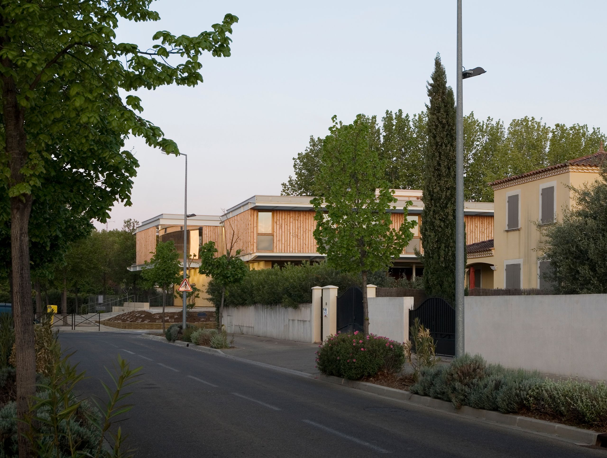 Vue de rue de l’école et centre de loisirs Jean Carrière à Nîmes, volumes en bois surélevés entre les platanes, toitures plates et façades rythmées verticalement.