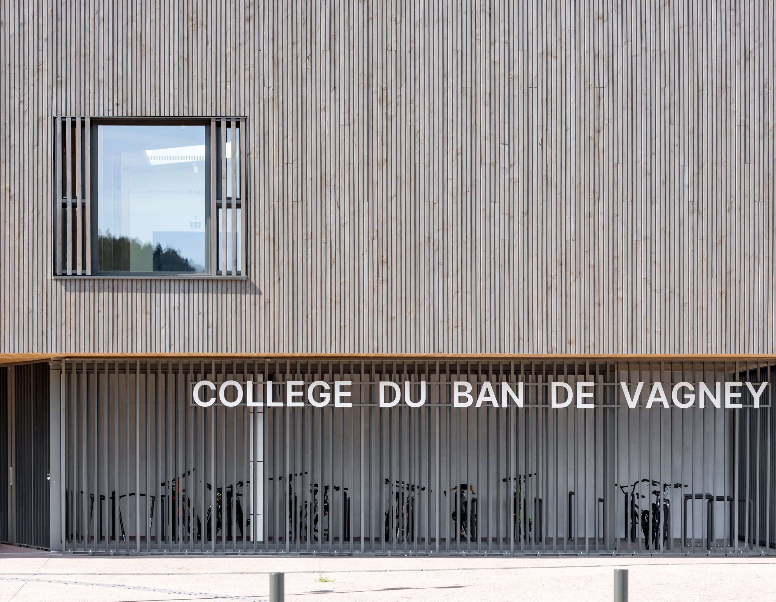 Façade bois du Collège du Ban à Vagney, bardage à claire-voie, grande baie vitrée et rez-de-chaussée ajouré servant d’abri vélos sous l’enseigne du collège.