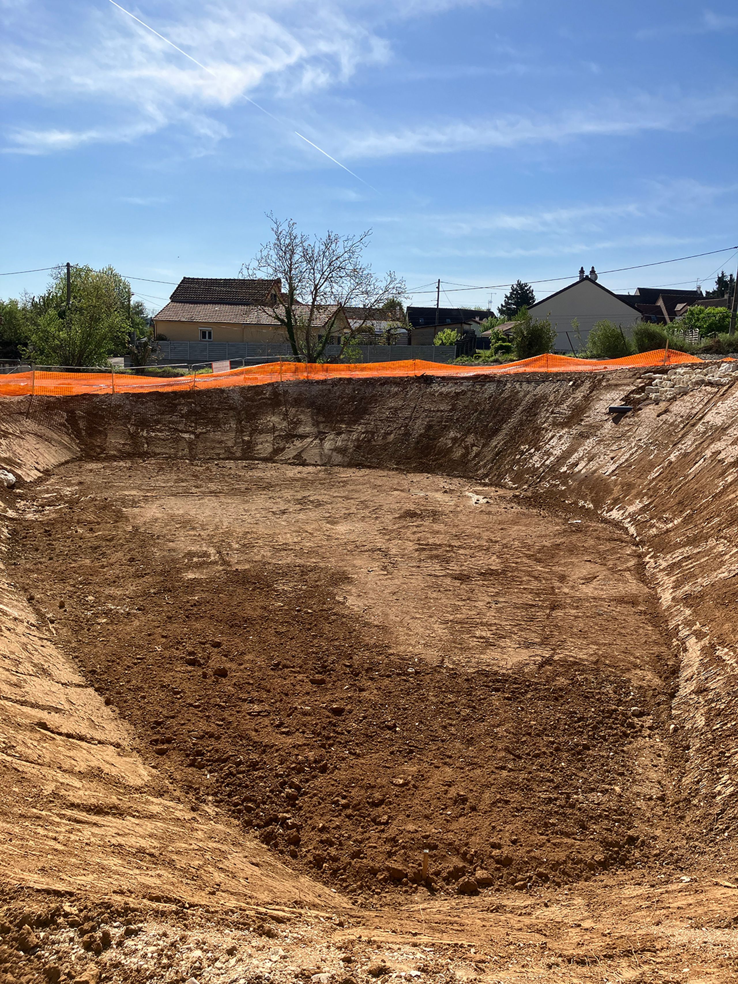 Excavated basin for the Auxerre nursing home, showing earthworks contained by orange safety fencing, with the existing residential context in the background.