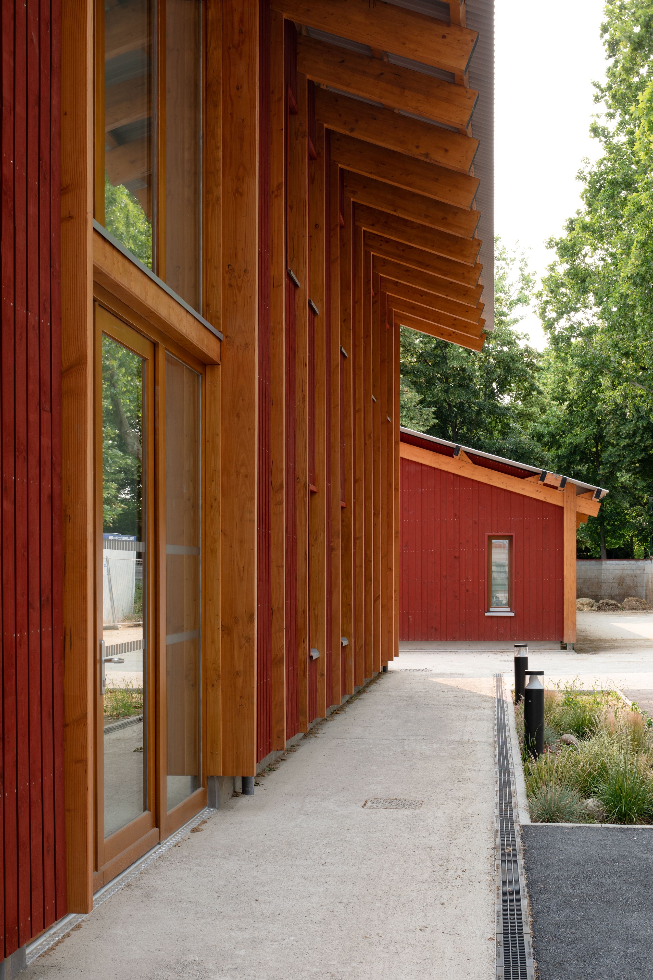 Façades en bois et bardage rouge de Falun du centre équestre de Gennevilliers, alignement de poteaux et auvent profond cadrant une circulation extérieure minérale.
