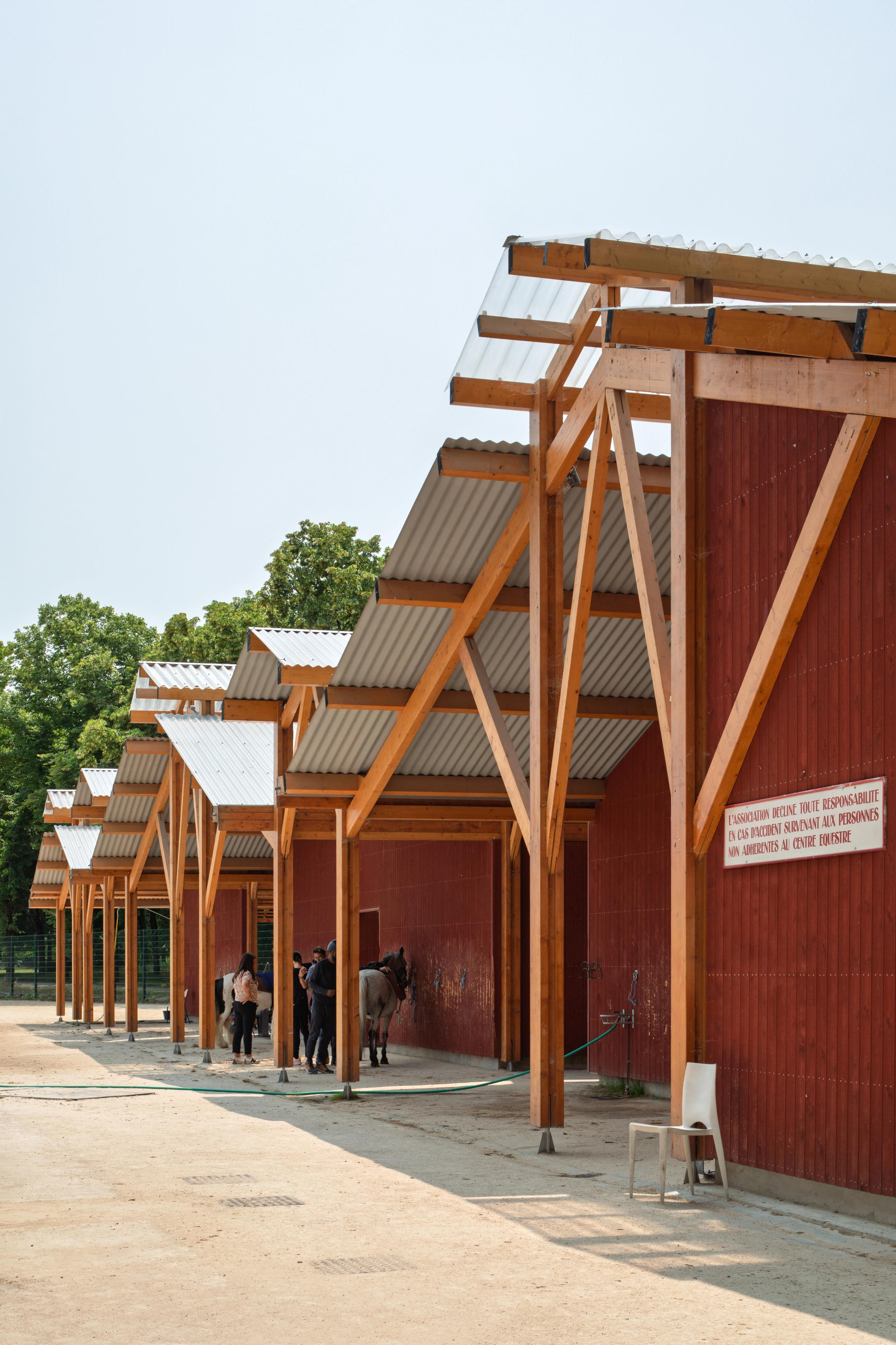 Timber-framed stables of the Gennevilliers equestrian center, with Falun red cladding and corrugated roofs forming a covered circulation along the arena.