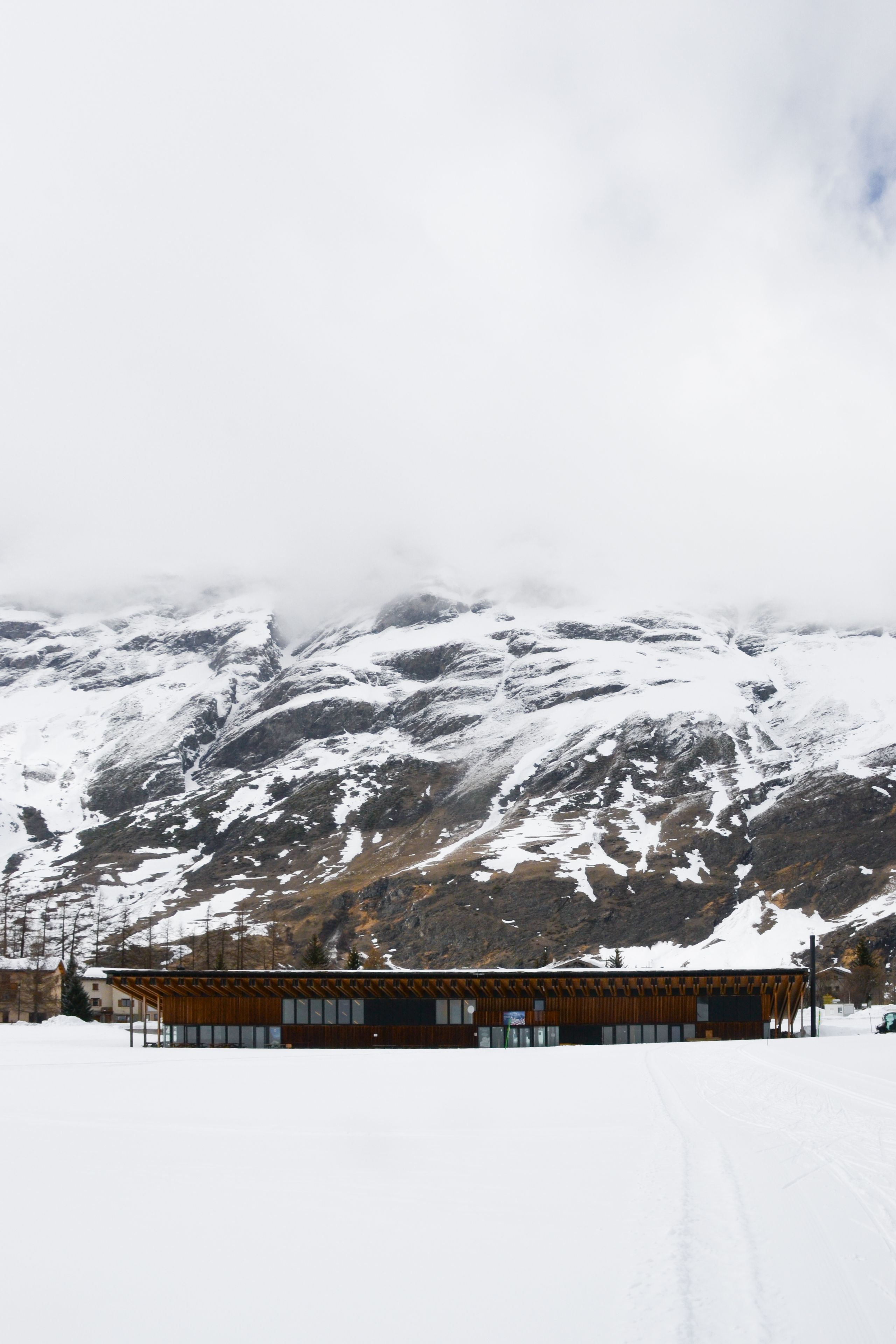 Espace nordique de Bessans : volume bas en bois et verre, toiture à pans inclinés, implanté en lisière des pistes de ski face au massif enneigé de Haute-Maurienne.