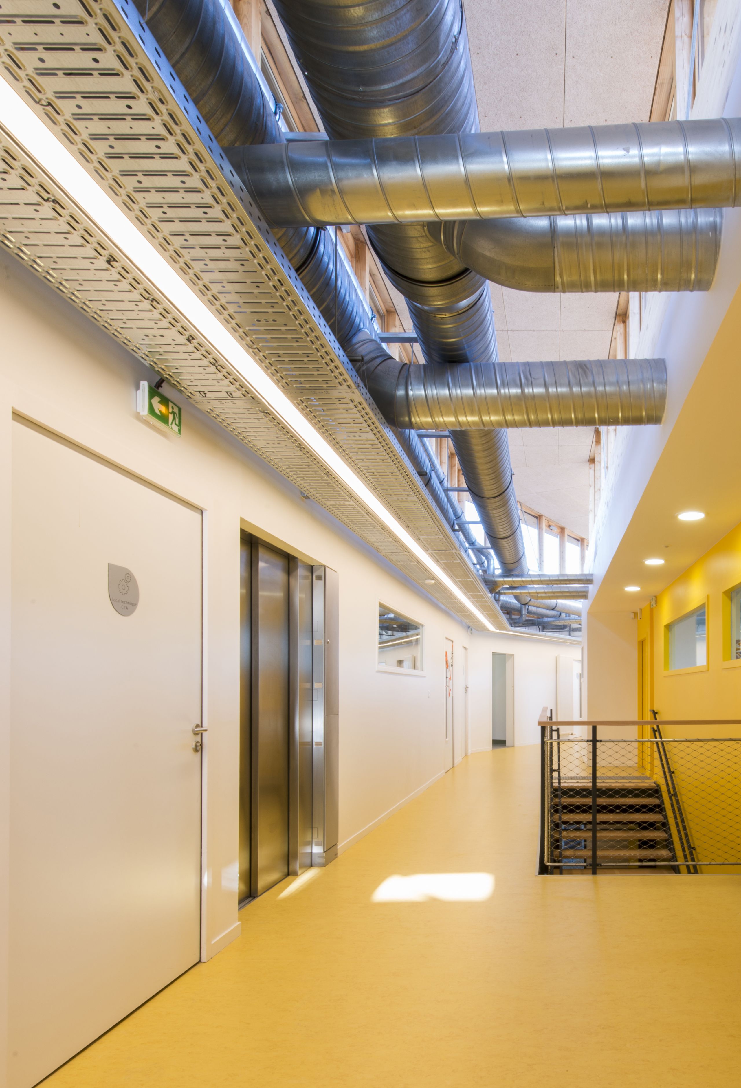 Circulation intérieure du centre de loisirs de Givors, couloir lumineux avec gaines techniques apparentes, noyau d’ascenseur et escalier métallique.