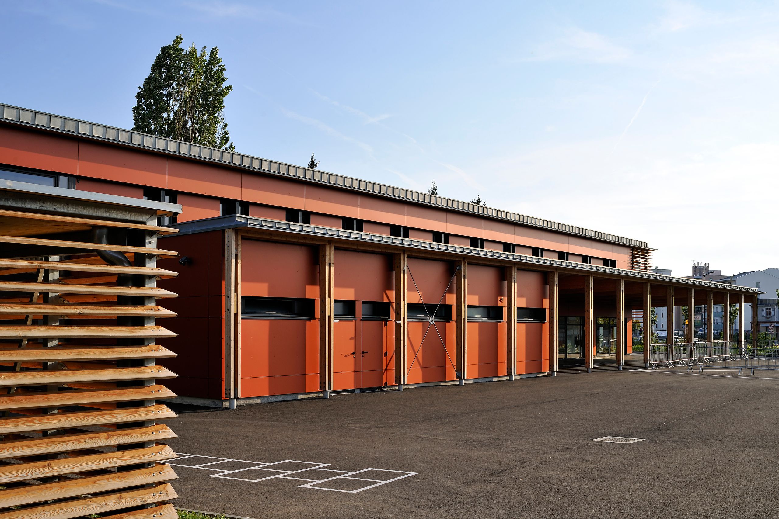 Façade du groupe scolaire Antoine de Saint-Exupéry à Annemasse, volume bas en panneaux orange, structure bois apparente et large auvent sur cour minérale.