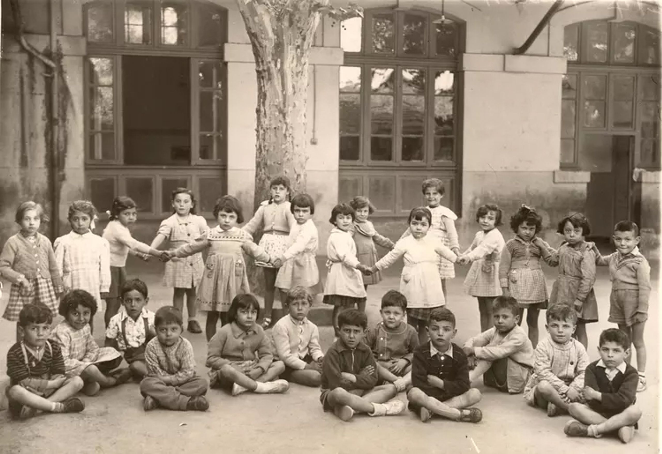 Photographie d’archive d’une cour d’école à Marseille : façade enduite à grandes baies vitrées bois, soubassement sombre et arbre central structurant l’espace.