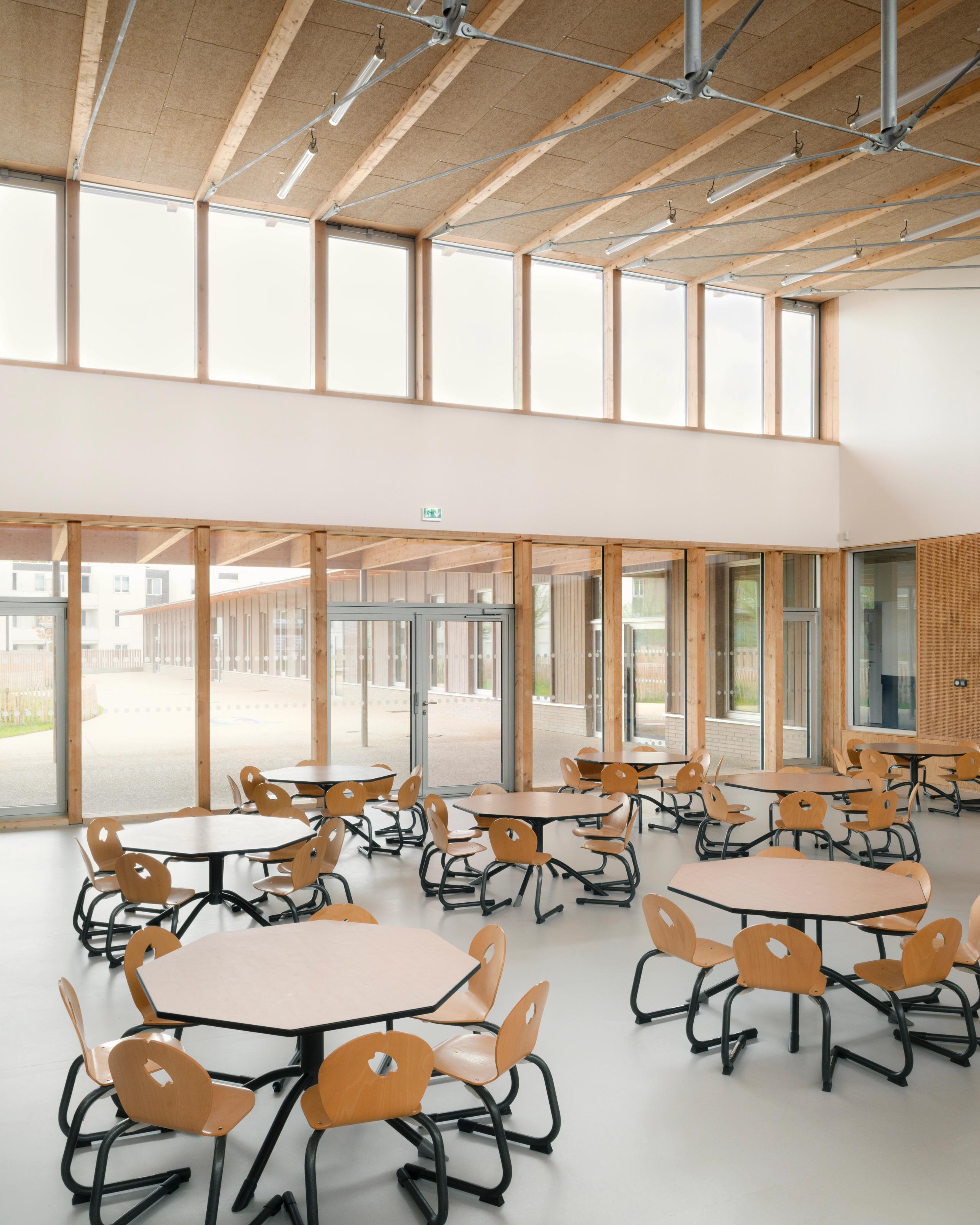 Cafeteria des groupes scolaires Jacqueline Auriol et Jean Jaurès au Bourget, vaste volume bois et verre, tables hexagonales face au préau couvert.