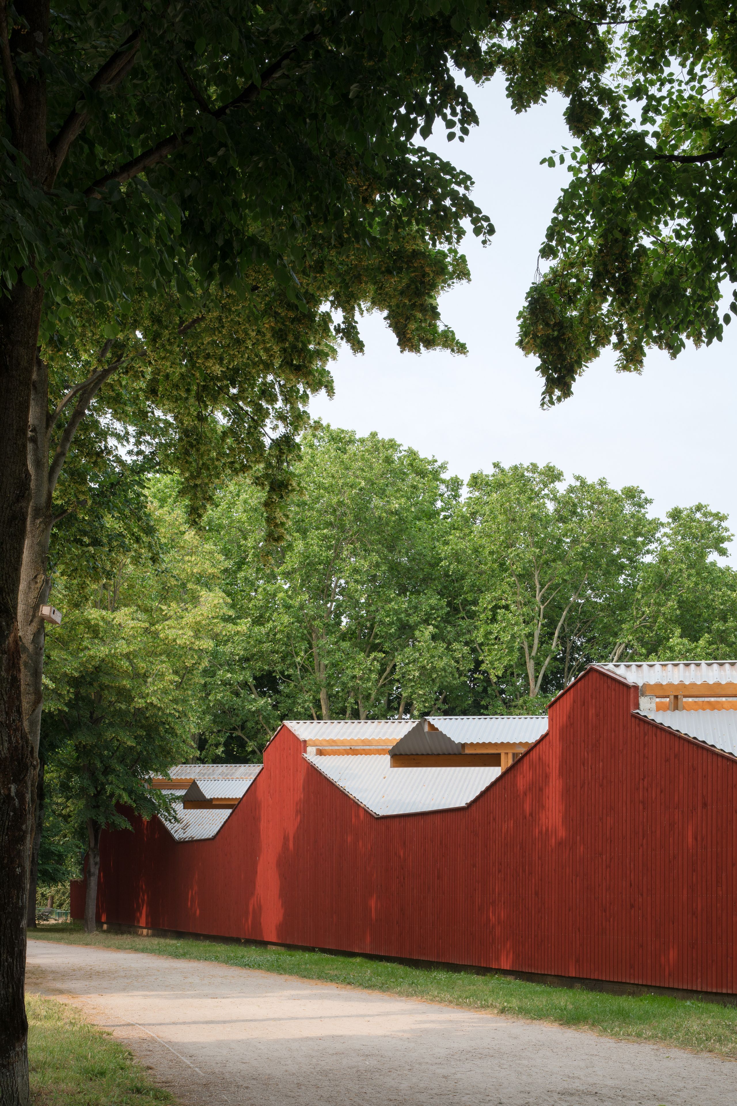 Centre équestre de Gennevilliers par Tectoniques : long volume en bois bardé de rouge de Falun, toitures métalliques plissées, en lisière d’un parc arboré.