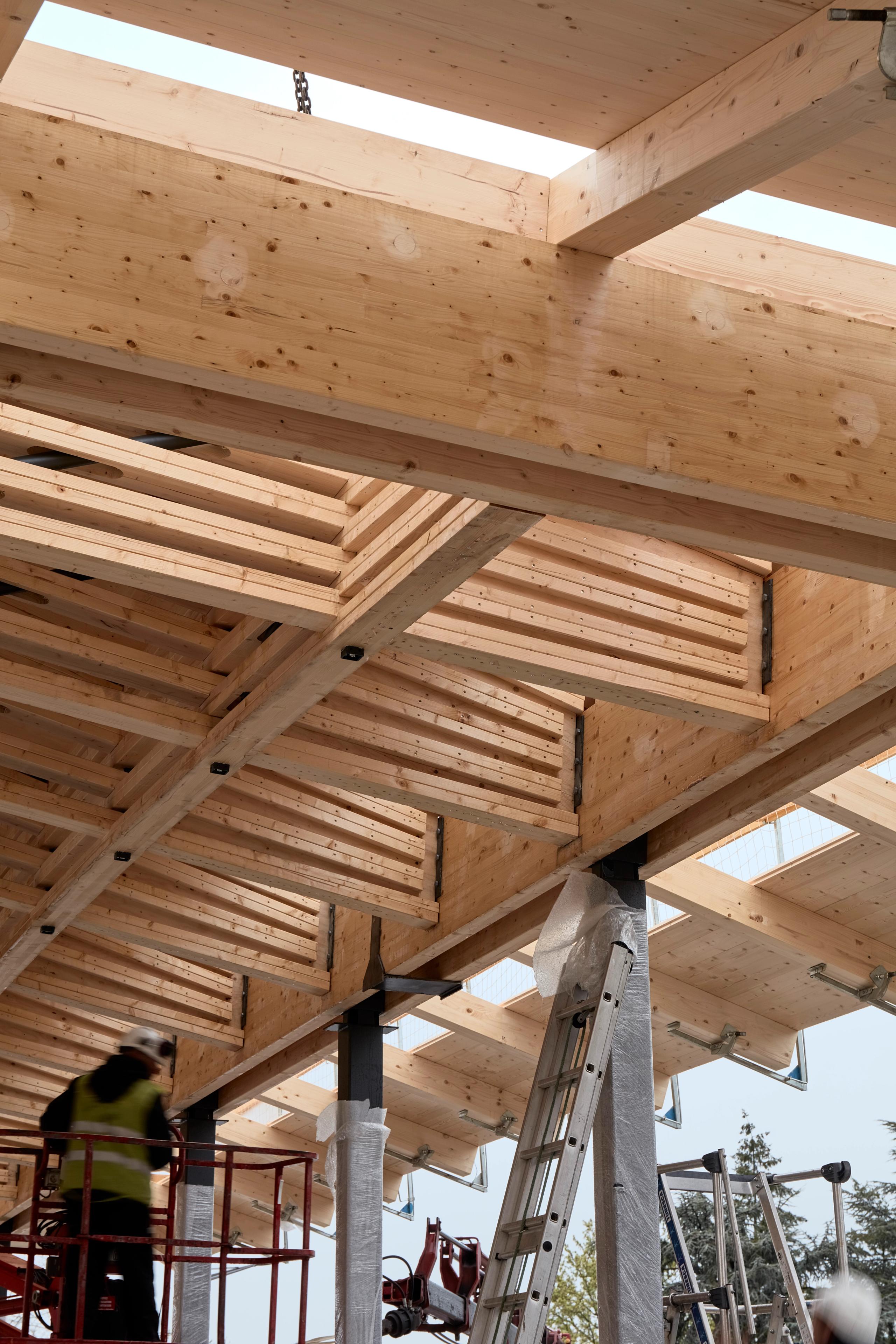 Chantier de la halle de marché de Marcy-l’Étoile : charpente en lamellé-collé formant une trame tridimensionnelle sur poteaux métalliques, vue depuis la place.