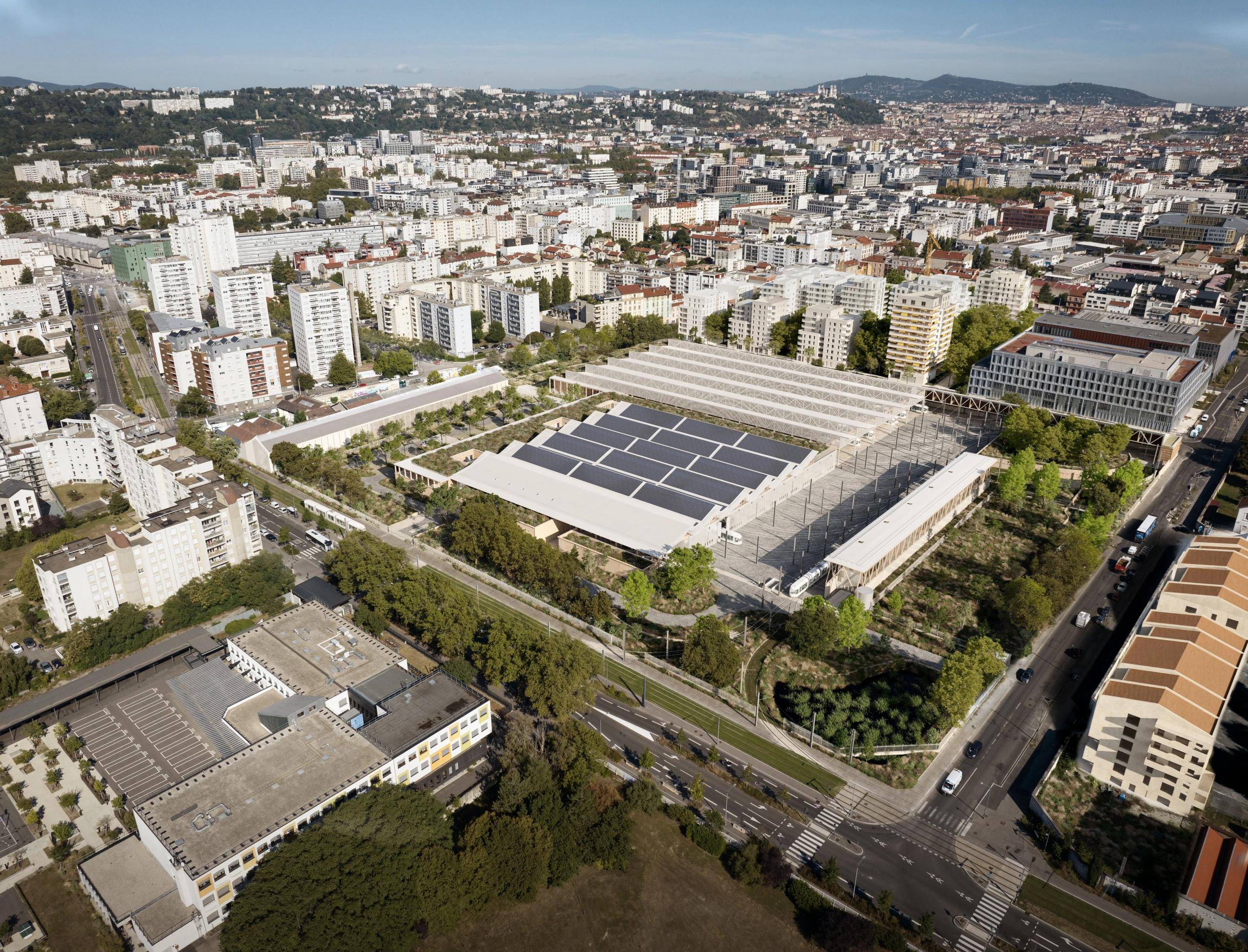 Vue aérienne du centre de maintenance de tramway à Lyon conçu par Tectoniques, implanté au cœur d’un tissu urbain dense : un vaste ensemble de halles basses et rationnelles, organisées en plateaux rectangulaires, dont les toitures plates sont largement couvertes de panneaux photovoltaïques et de structures métalliques légères, encadré par des voiries, des lignes de tramway et des barres de logements collectifs, avec une trame paysagère d’alignements d’arbres et d’espaces verts structurant la parcelle.