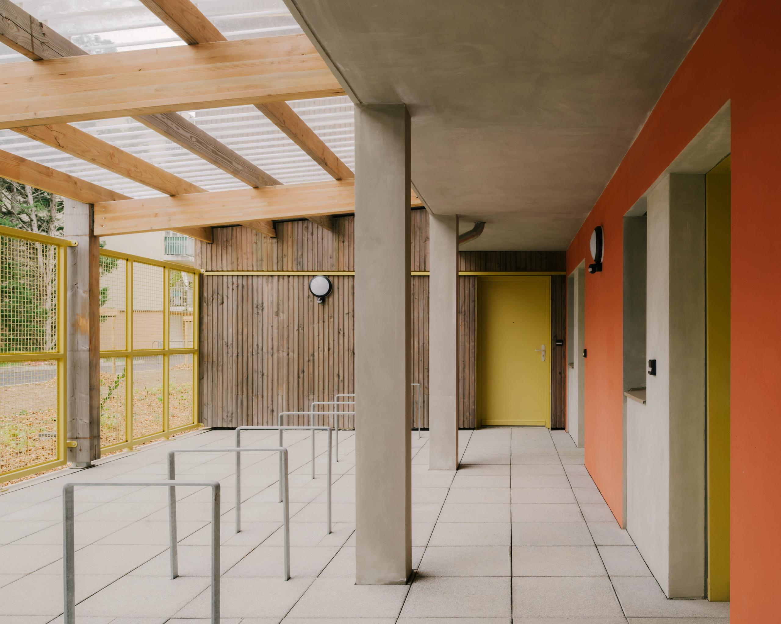Coursive couverte des 84 logements à Trévoux, structure bois-béton, bardage bois, poteaux béton, garde-corps métalliques et menuiseries jaunes et orange.