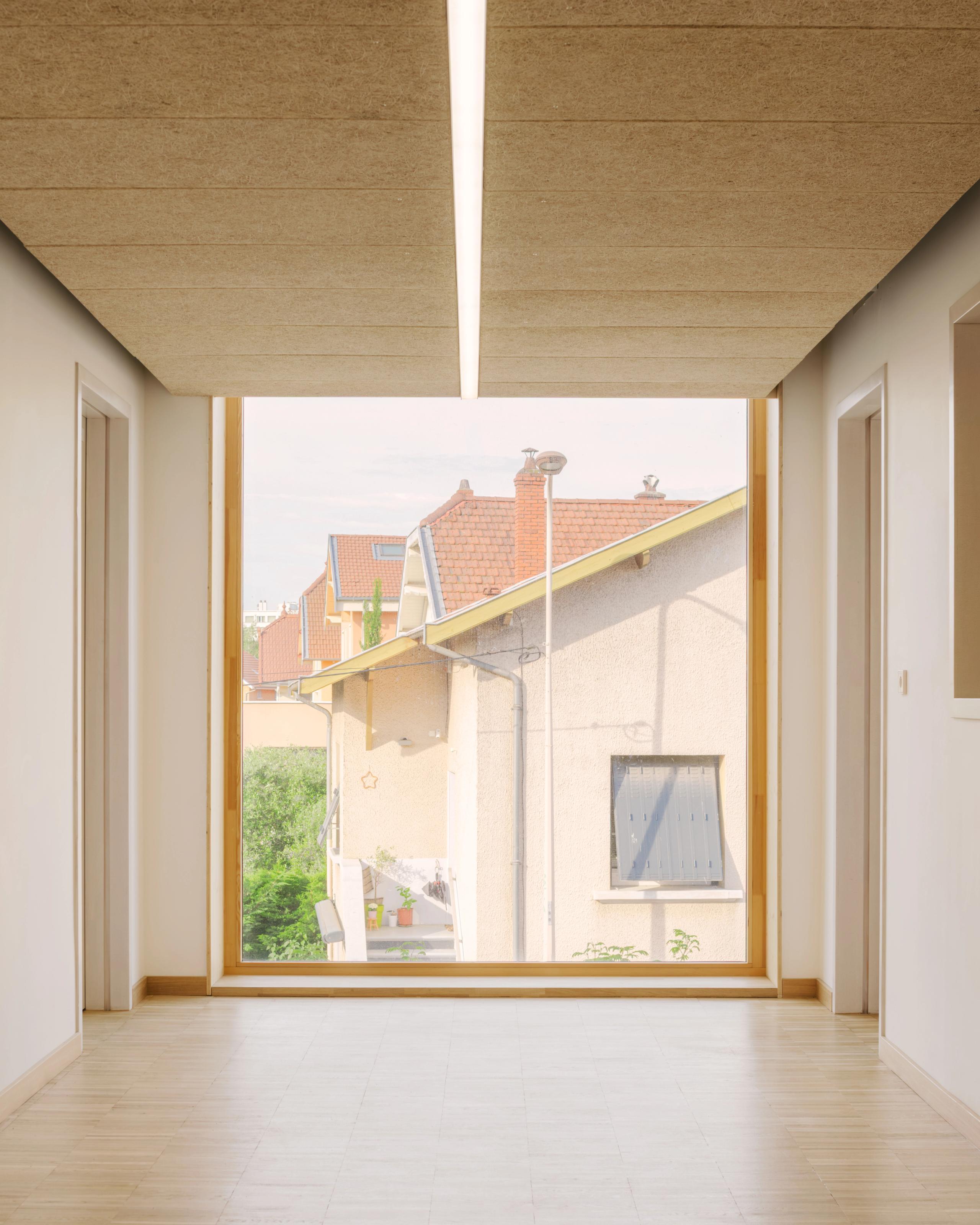 Couloir du groupe scolaire Kennedy à Lyon, éclairé par un plafond bois et un grand châssis vitré cadrant le tissu faubourien environnant.
