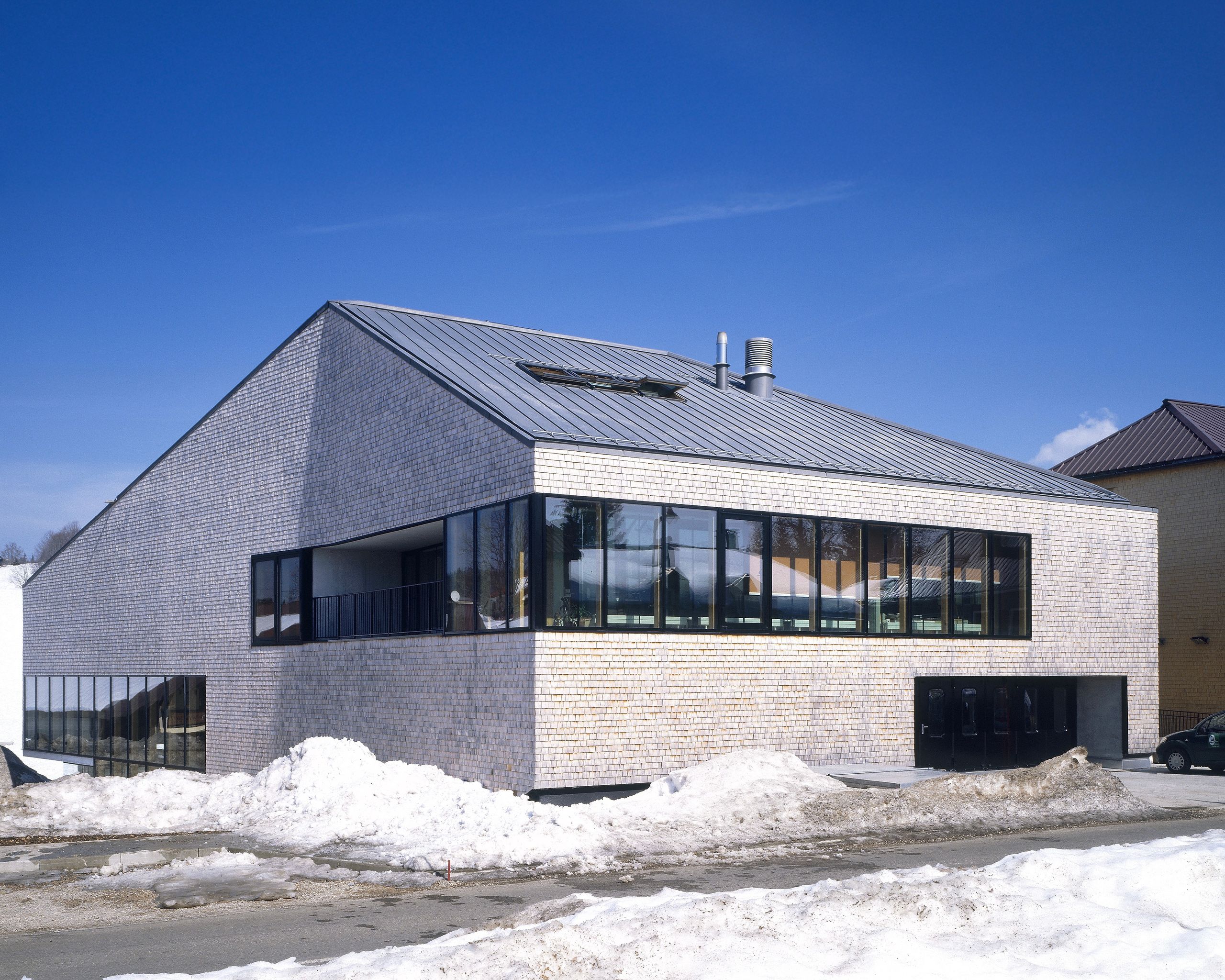 Extension en tavaillons d’épicéa de la Maison du Parc du Haut-Jura à Lajoux, volume compact à double peau vitrée, large bande de fenêtres sur rue enneigée.
