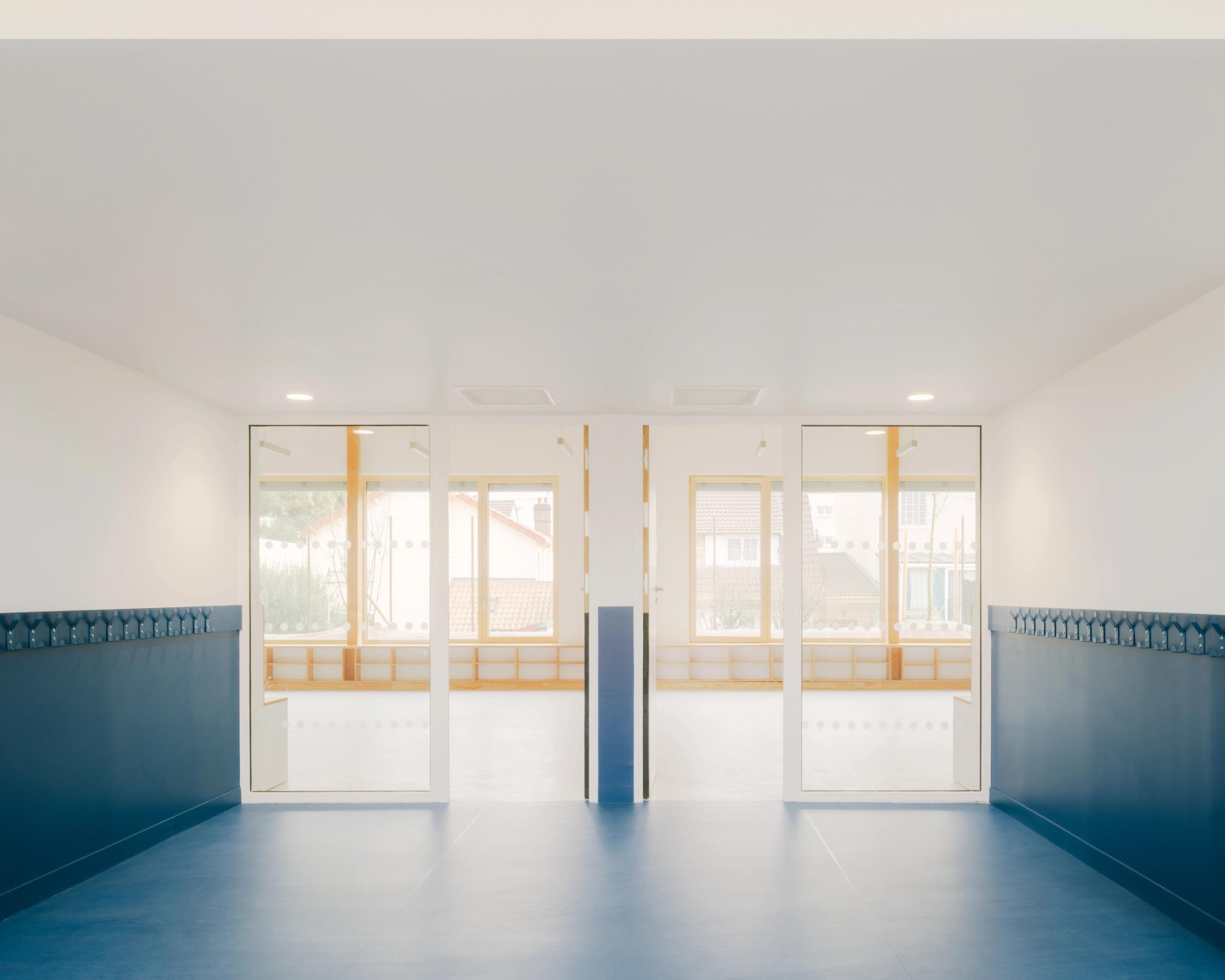 Intérieur des groupes scolaires Jacqueline Auriol et Jean Jaurès au Bourget, couloir bleu structuré par de larges baies vitrées et menuiseries bois claires.
