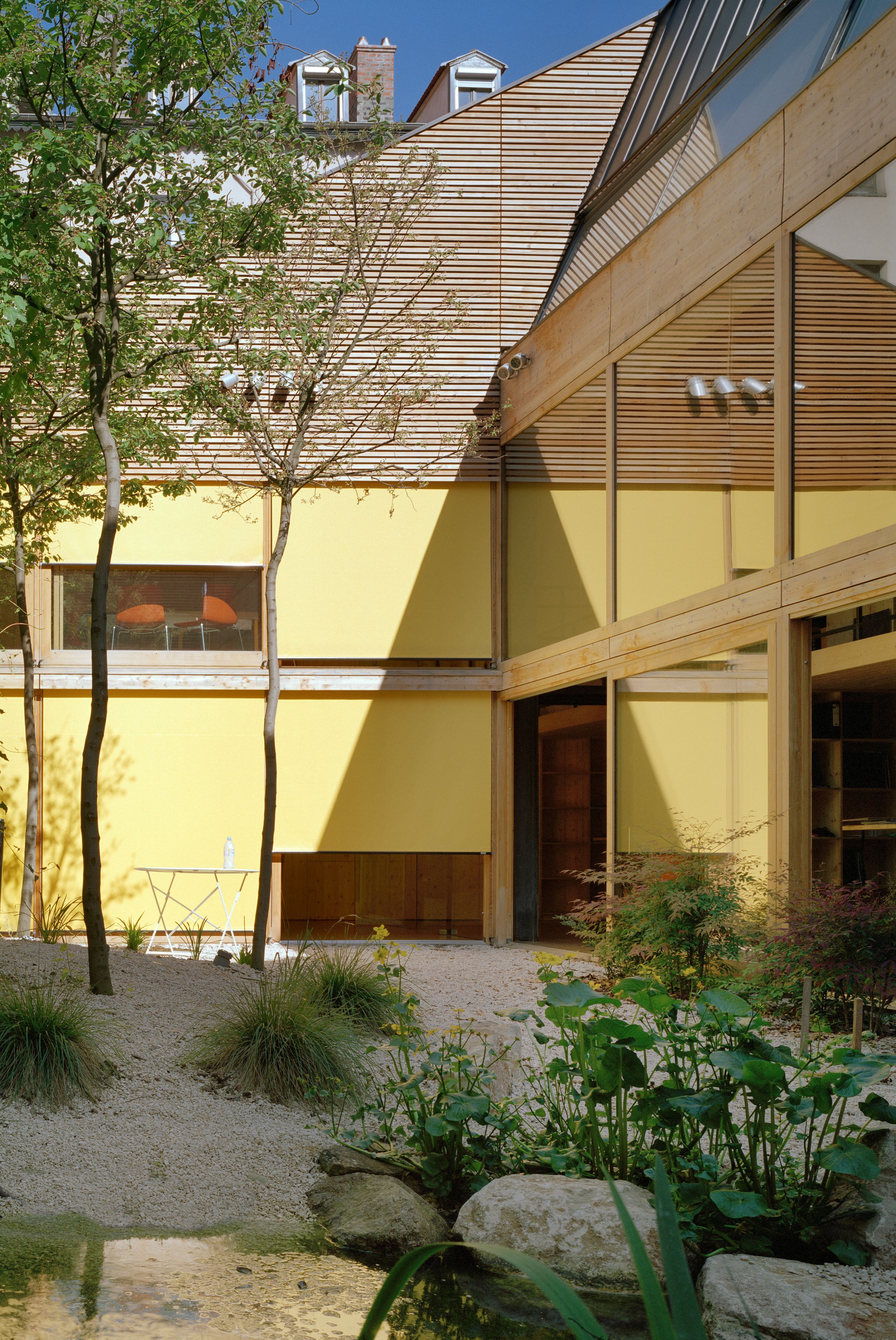 Patio calme de la réhabilitation d’un local d’activité à Lyon par Tectoniques, façade bois et enduit jaune, grandes baies vitrées sur jardin minéral avec bassin.