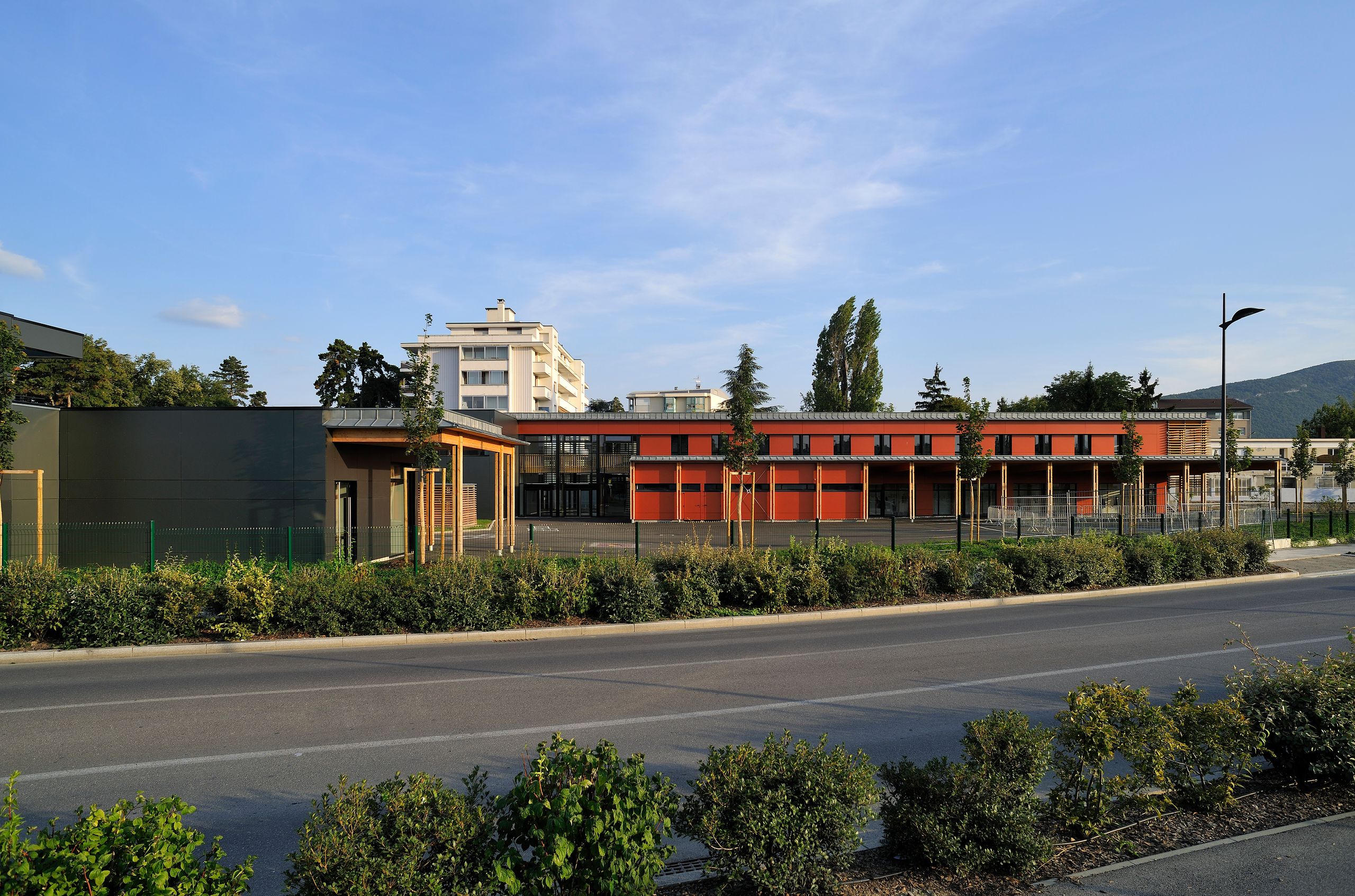 Façade du groupe scolaire Antoine de Saint-Exupéry à Annemasse, volumes bas en bois et panneaux orange, large préau vitré sur rue paysagée.