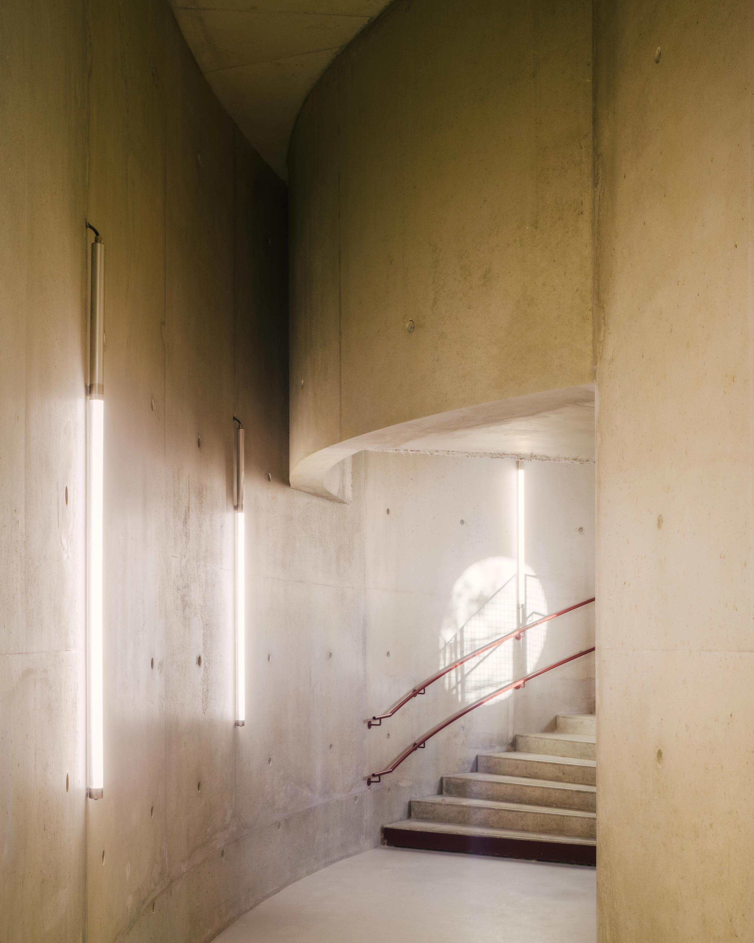 Circulation intérieure du groupe scolaire Dominique Frelaut à Colombes, escalier en béton courbe, garde-corps métalliques rouges et éclairage linéaire mural.