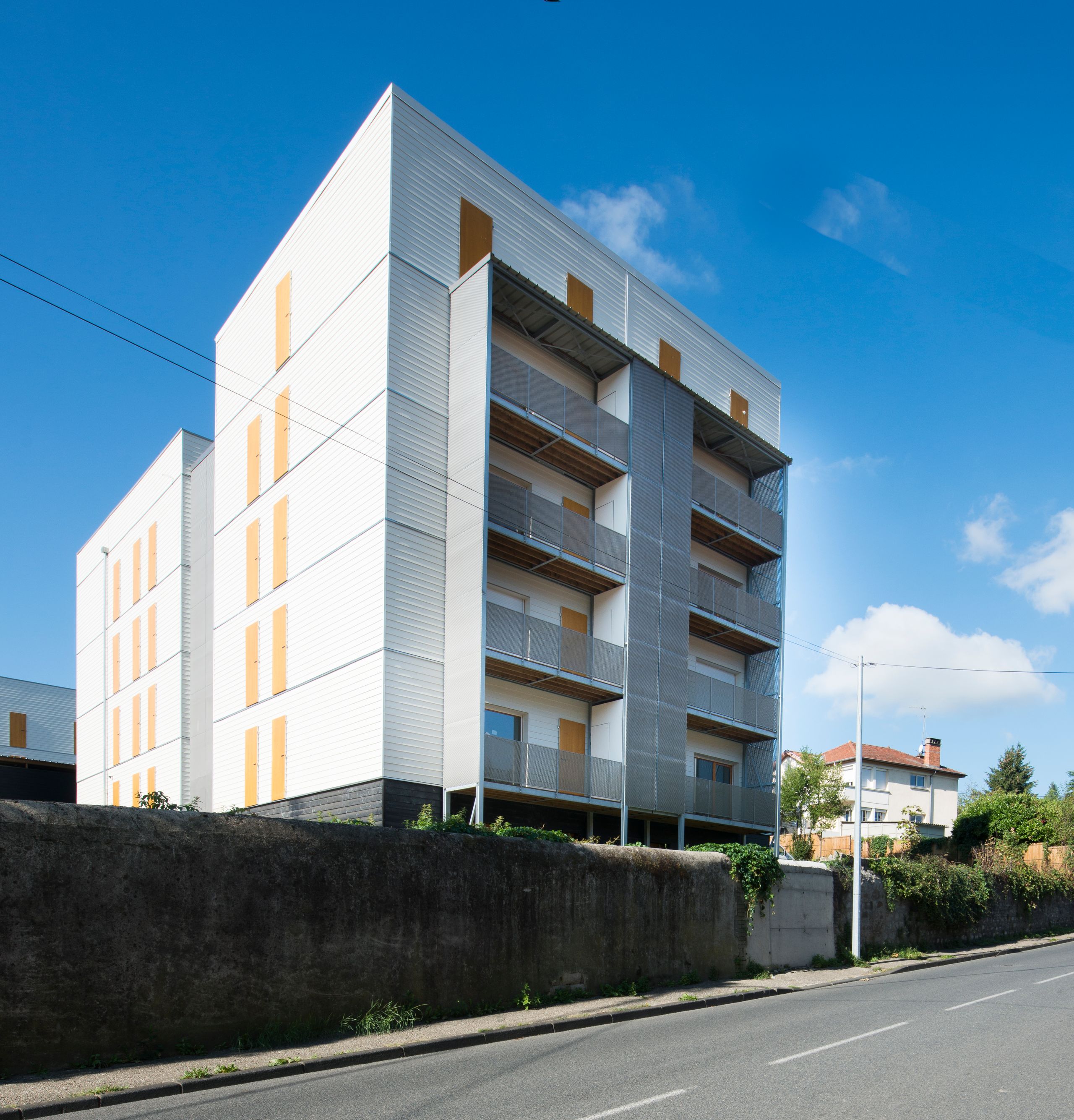 Tectoniques, 60 logements à Rive-de-Gier : petit collectif en façade métallique claire, balcons filants et percements bois, implanté en surplomb d’une rue en pente.