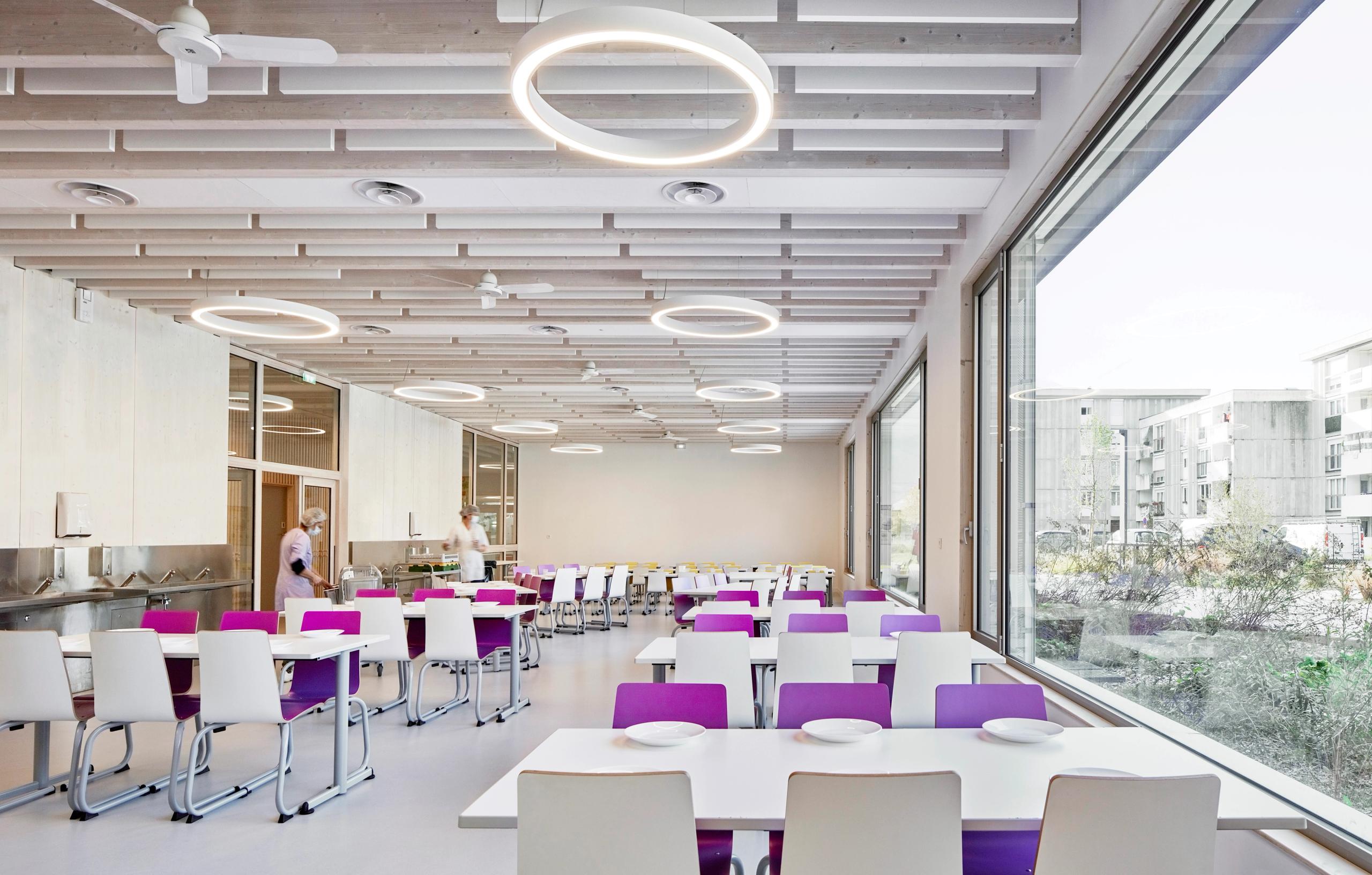 Cafeteria de la Maison de l’Enfance Simone Veil à Albertville, vaste volume en bois apparent, luminaires circulaires et façade largement vitrée sur le quartier.