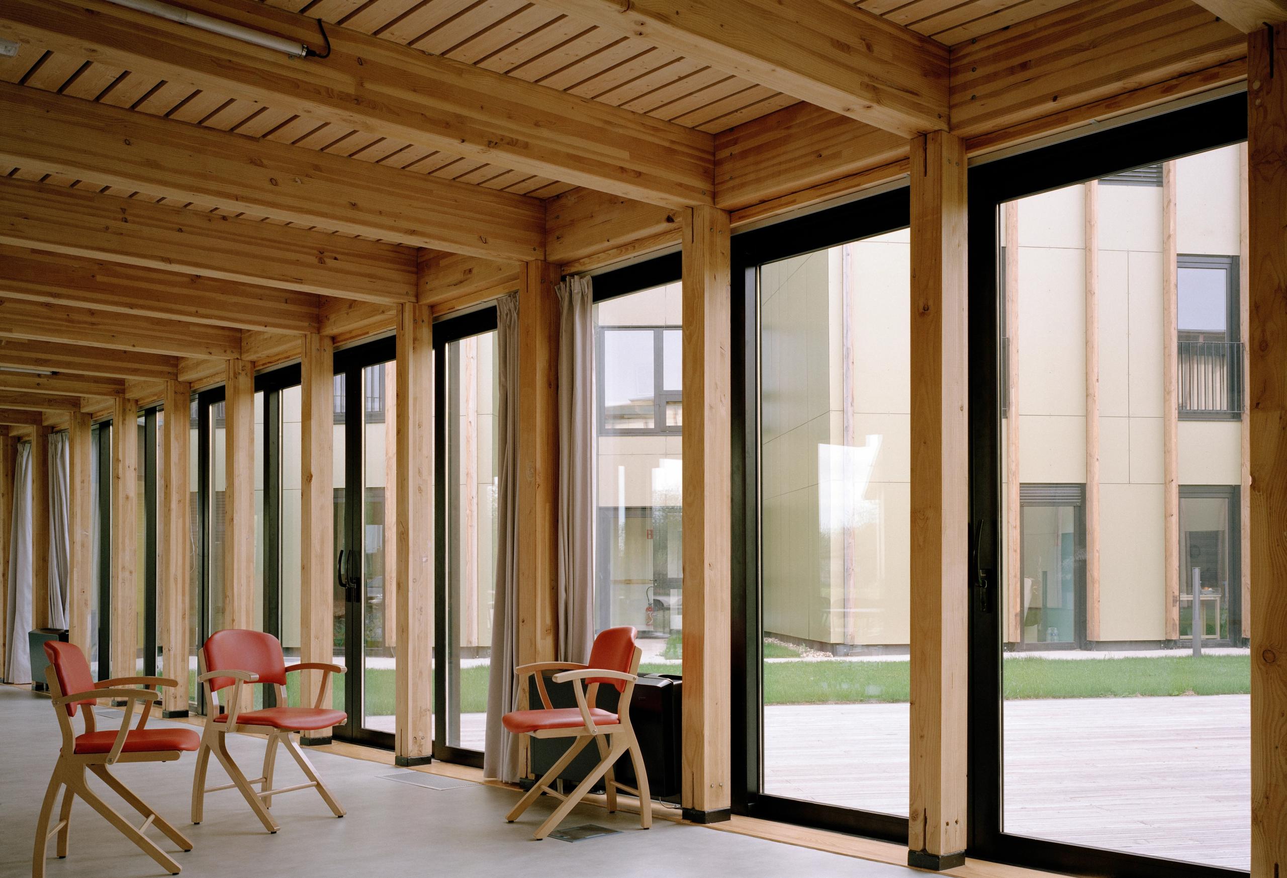 Galerie vitrée en structure bois de l’EHPAD de Mervans, ouvrant sur le jardin clos, avec poteaux, chevrons apparents et assises pour les unités de vie.