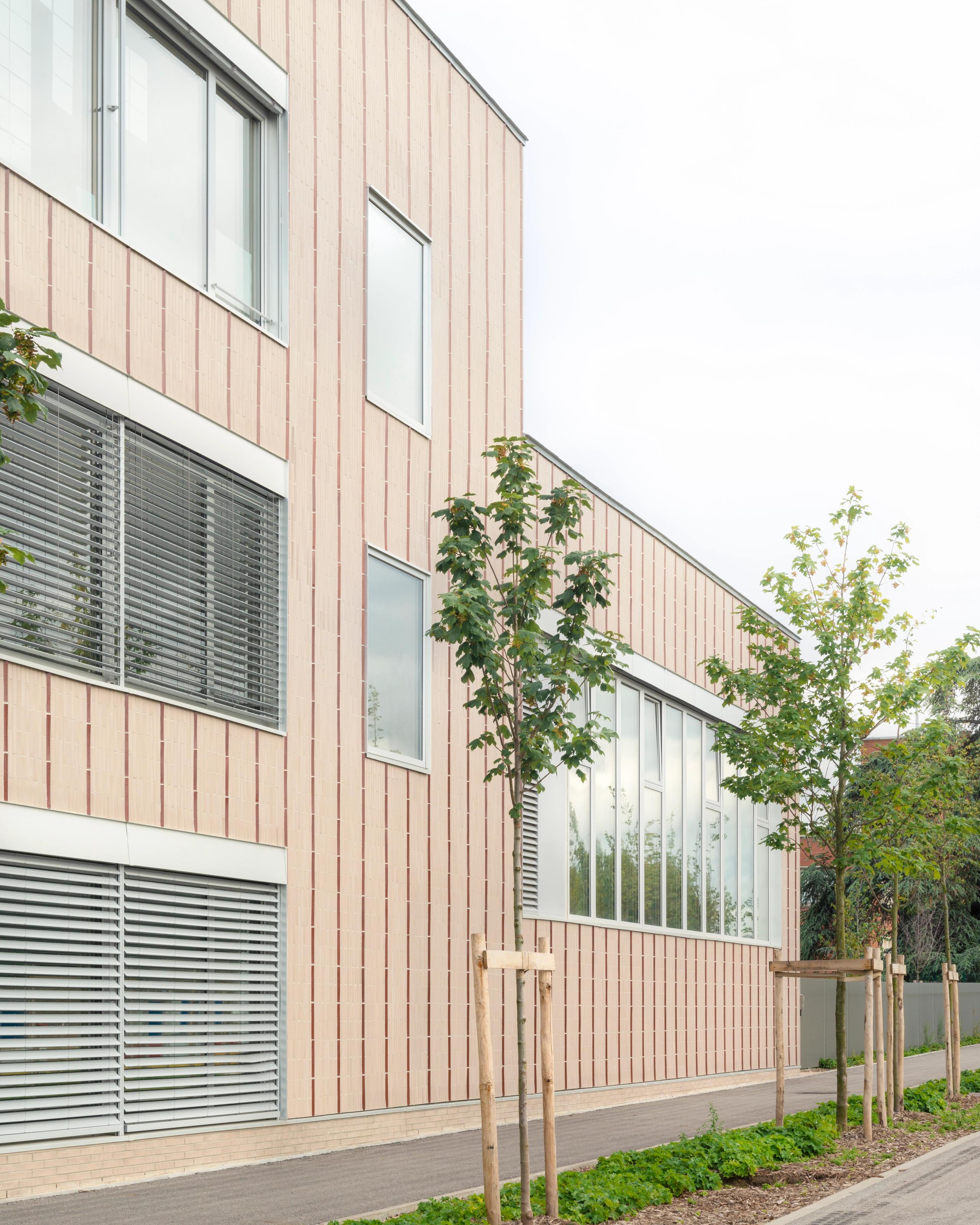 Façade en U des groupes scolaires Jacqueline Auriol et Jean Jaurès au Bourget, bardage en terre cuite claire, grandes baies vitrées et alignement d’arbres jeunes.