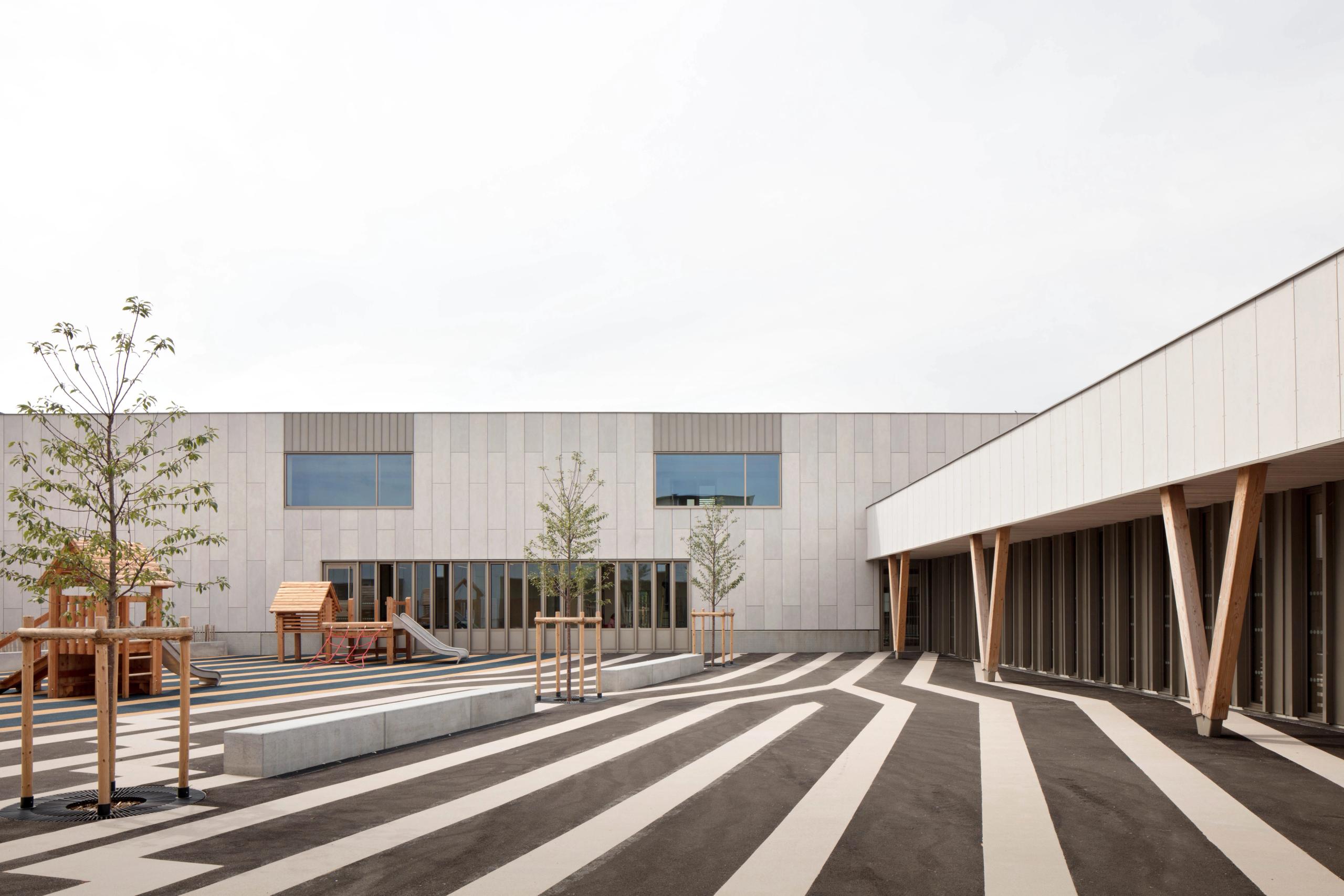 Cour sud du groupe scolaire Denis Mukwege à Melun, façade minérale rythmée, préau bois, sol rayé noir et beige structurant les circulations et jeux.