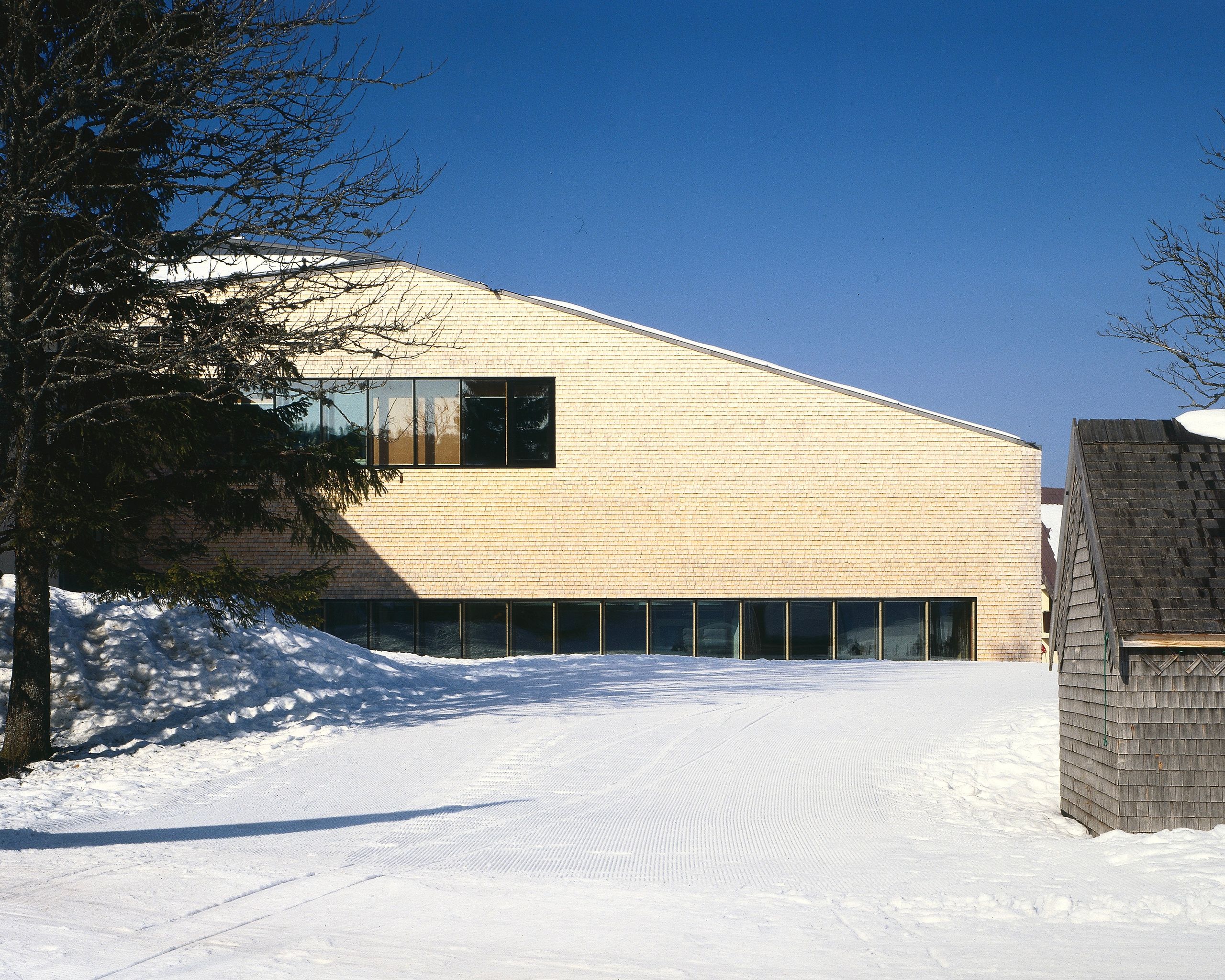 Extension en tavaillons d’épicéa de la Maison du Parc du Haut-Jura à Lajoux, volume compact et étiré, larges baies vitrées sur paysage enneigé.