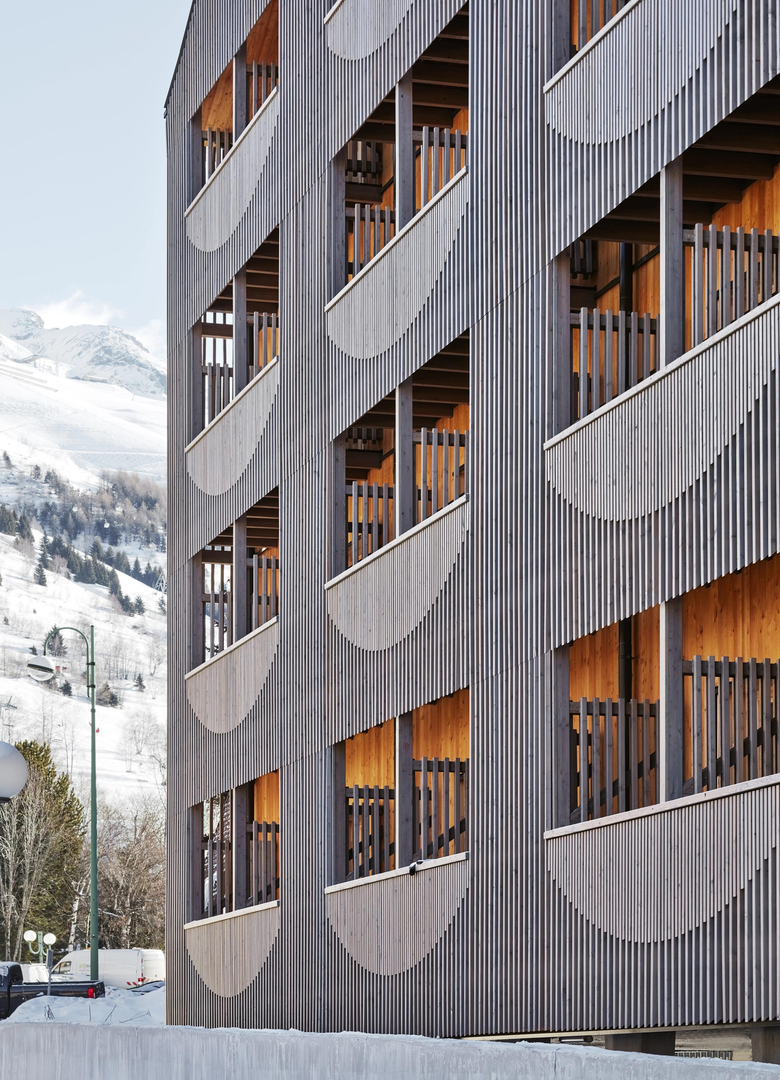 Façade bois à claire-voie de l’auberge de jeunesse réhabilitée aux Deux Alpes, balcons rythmés par une double peau ajourée face au paysage alpin enneigé.