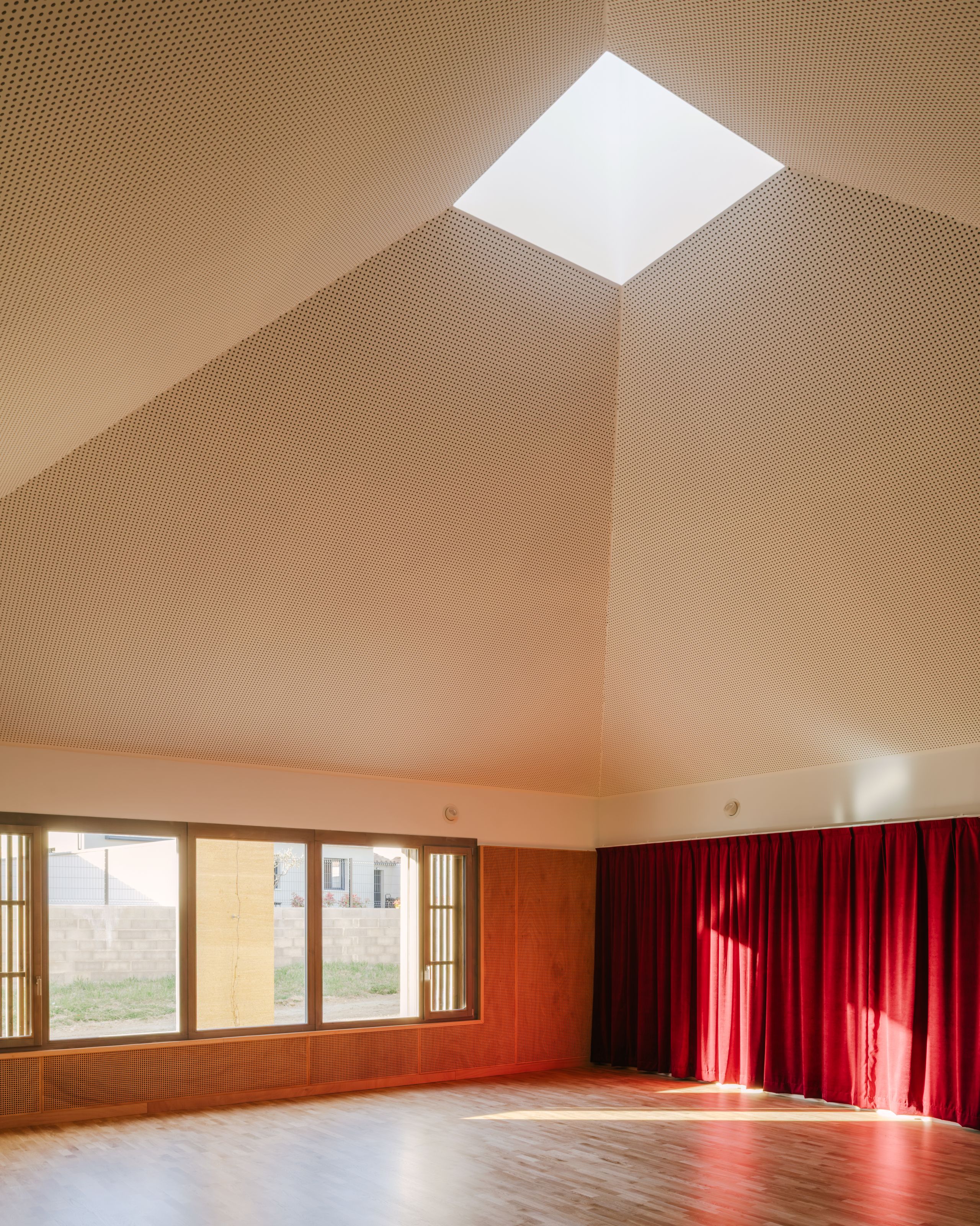 Salle du pôle culture et jeunesse à Vaison-la-Romaine, volume haut en bois avec plafond perforé, grand puits de lumière central et façade vitrée sur le lotissement.