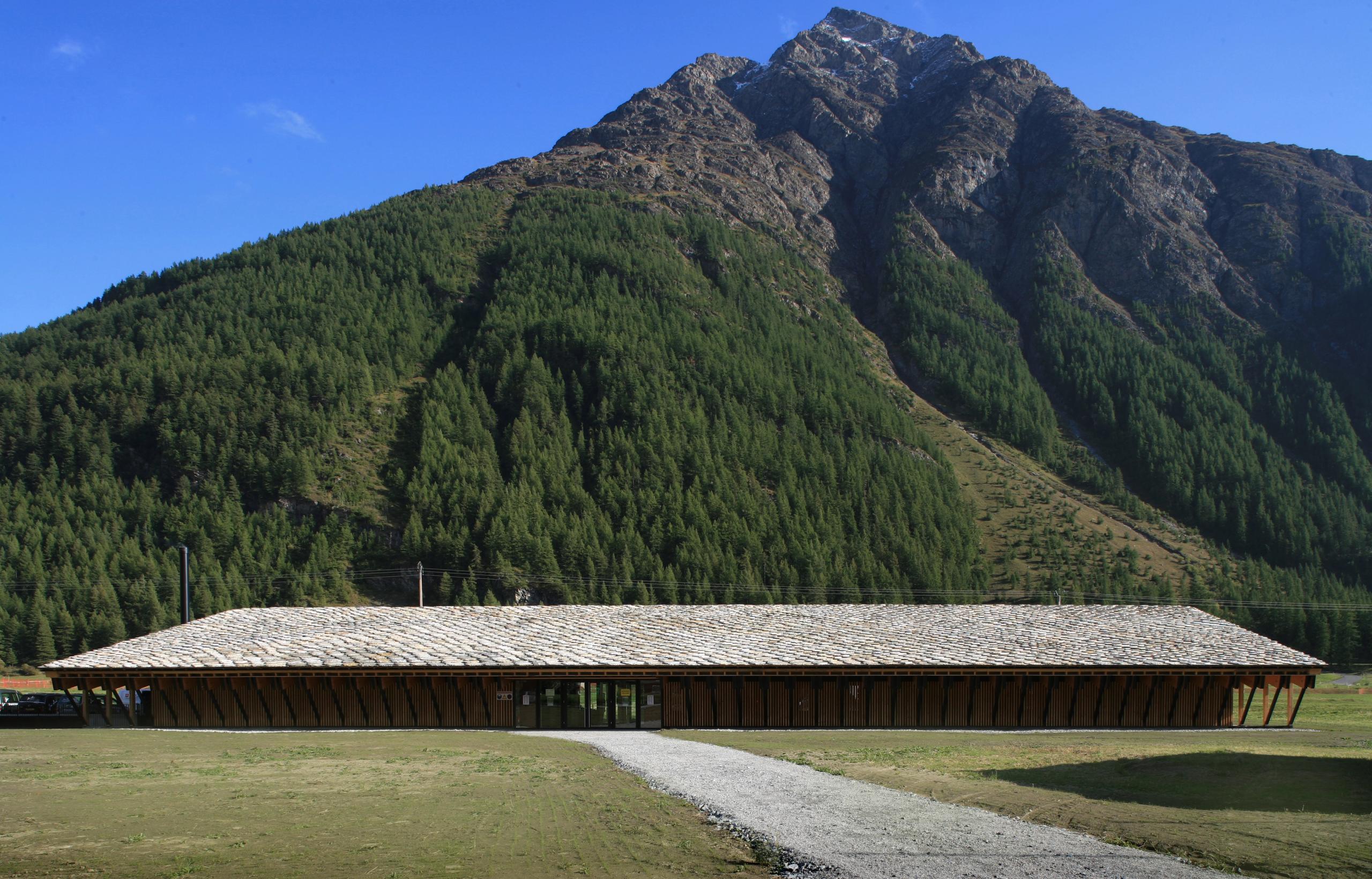 Espace nordique de Bessans : volume bas en bois et toiture en lauze, implanté face aux pistes, en pied de versant boisé de Haute-Maurienne.