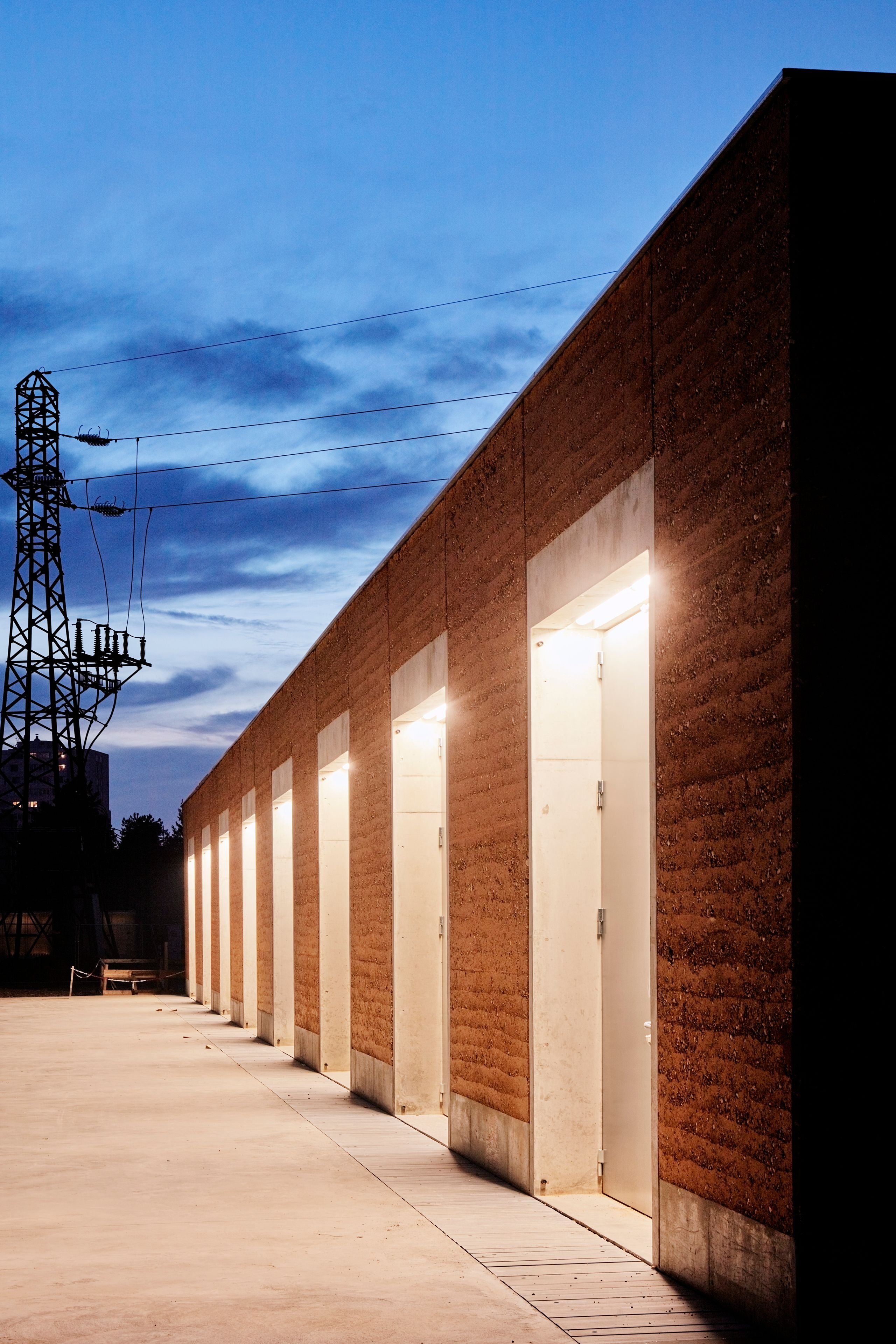 Poste source de Cusset à Villeurbanne : volume linéaire en pisé et béton brut, portes techniques éclairées, parvis minéral, pylône électrique en arrière-plan.