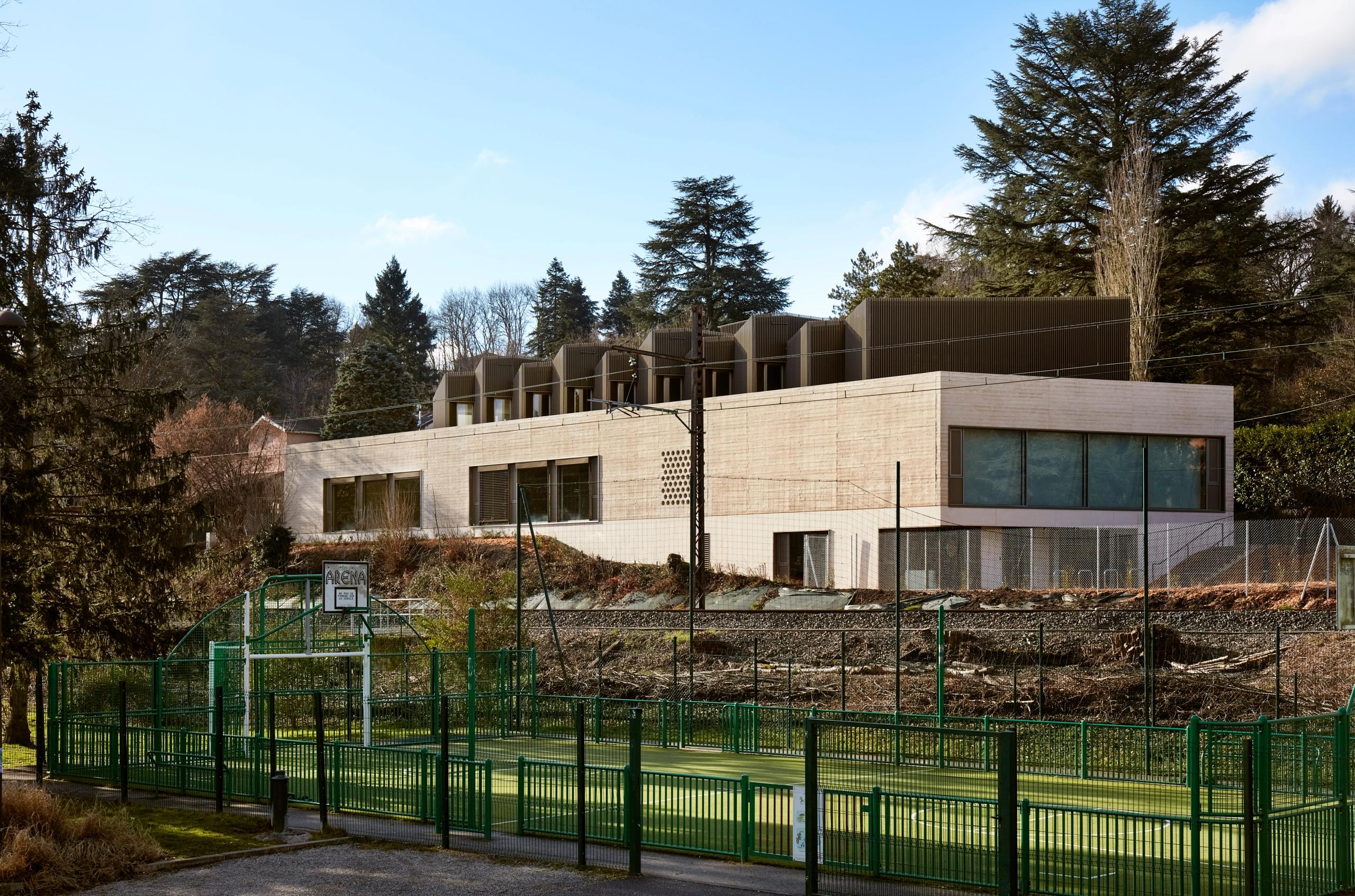 Vue de la Maison des Arts à Charbonnières-les-Bains : volume en béton en gradins dans la pente, couronnement musical en bois et métal perforé, surplombant un terrain de sport.