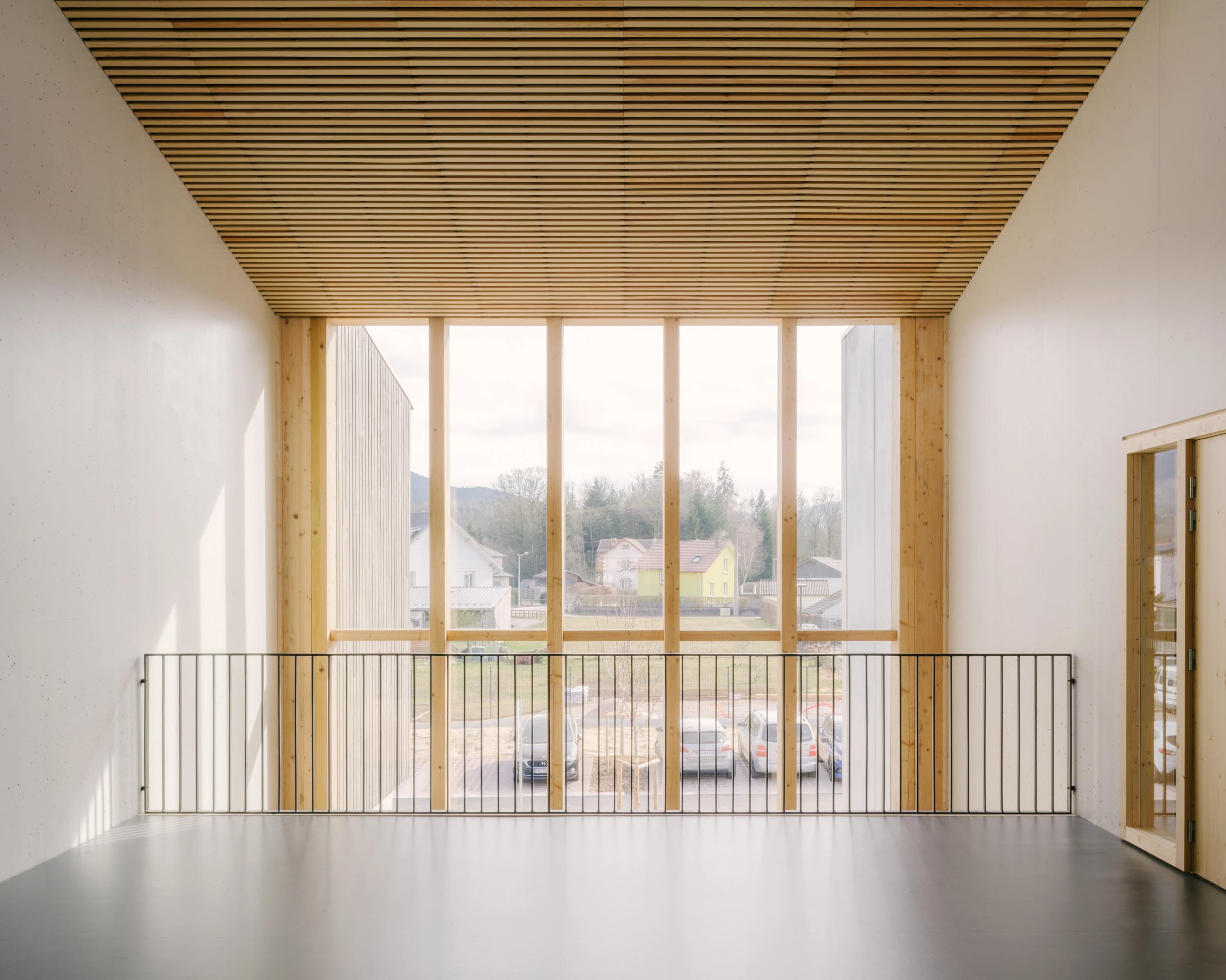 Vue intérieure du Collège du Ban à Vagney : rue intérieure en double hauteur, structure bois apparente, garde-corps métallique et large baie vitrée sur le paysage.