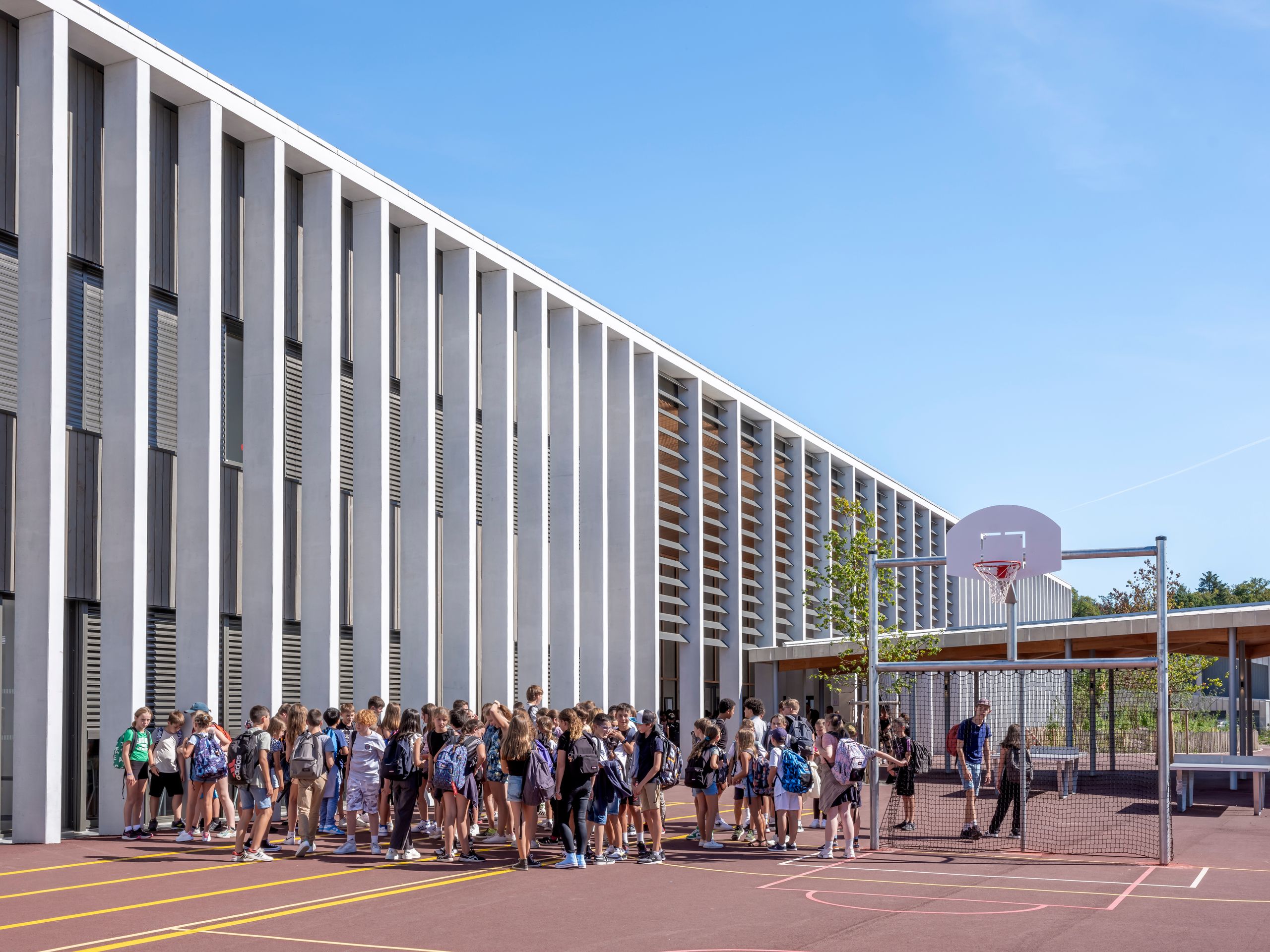 Vue de la cour du Collège du Ban à Vagney : façades en peigne à portique rythmé de poteaux béton et remplissages bois, préau et aire sportive occupée par les élèves.
