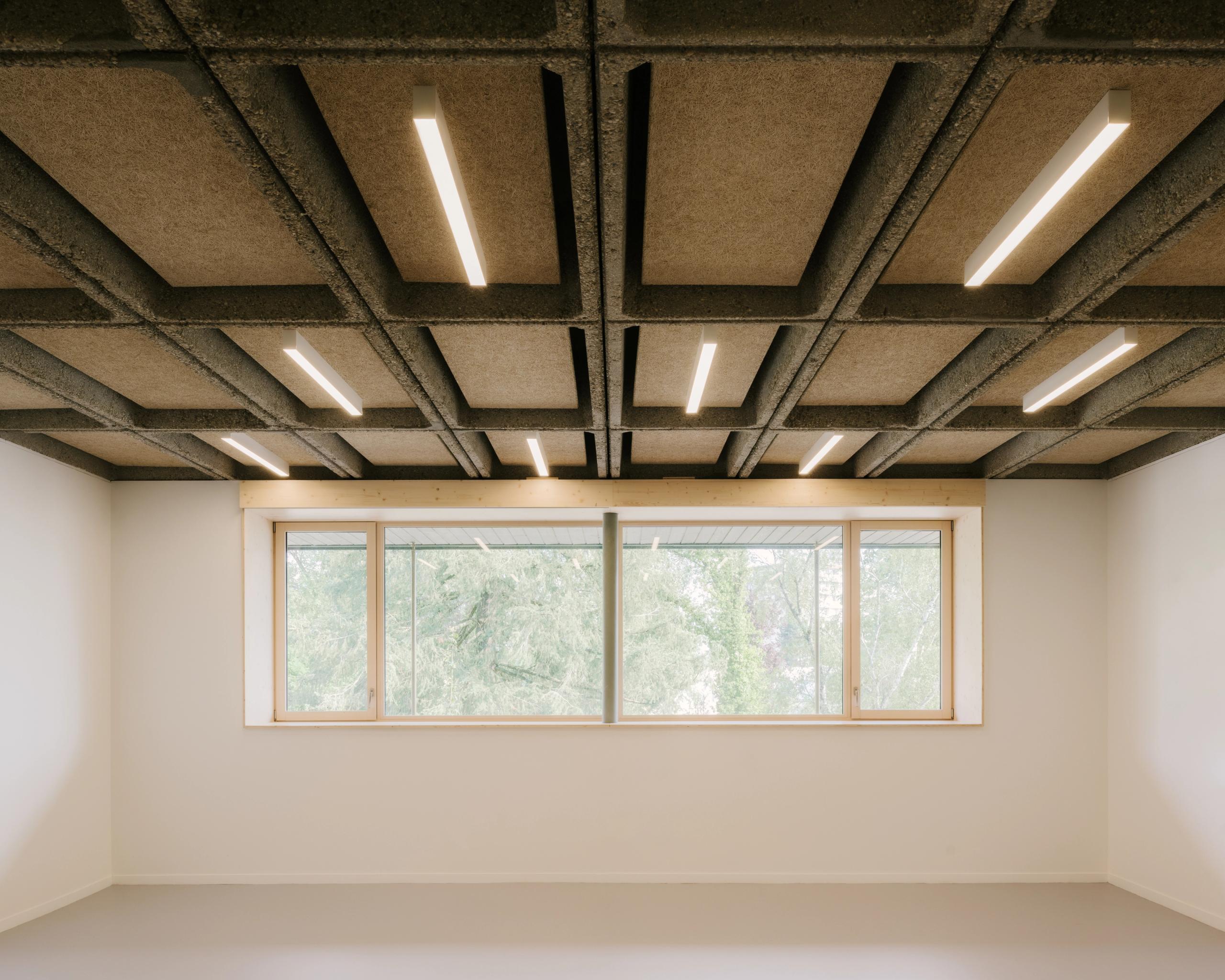 Salle du collège Maryse Bastié à Dole, montrant plafond caisson en béton apparent avec luminaires linéaires et grande baie vitrée bois sur paysage arboré.