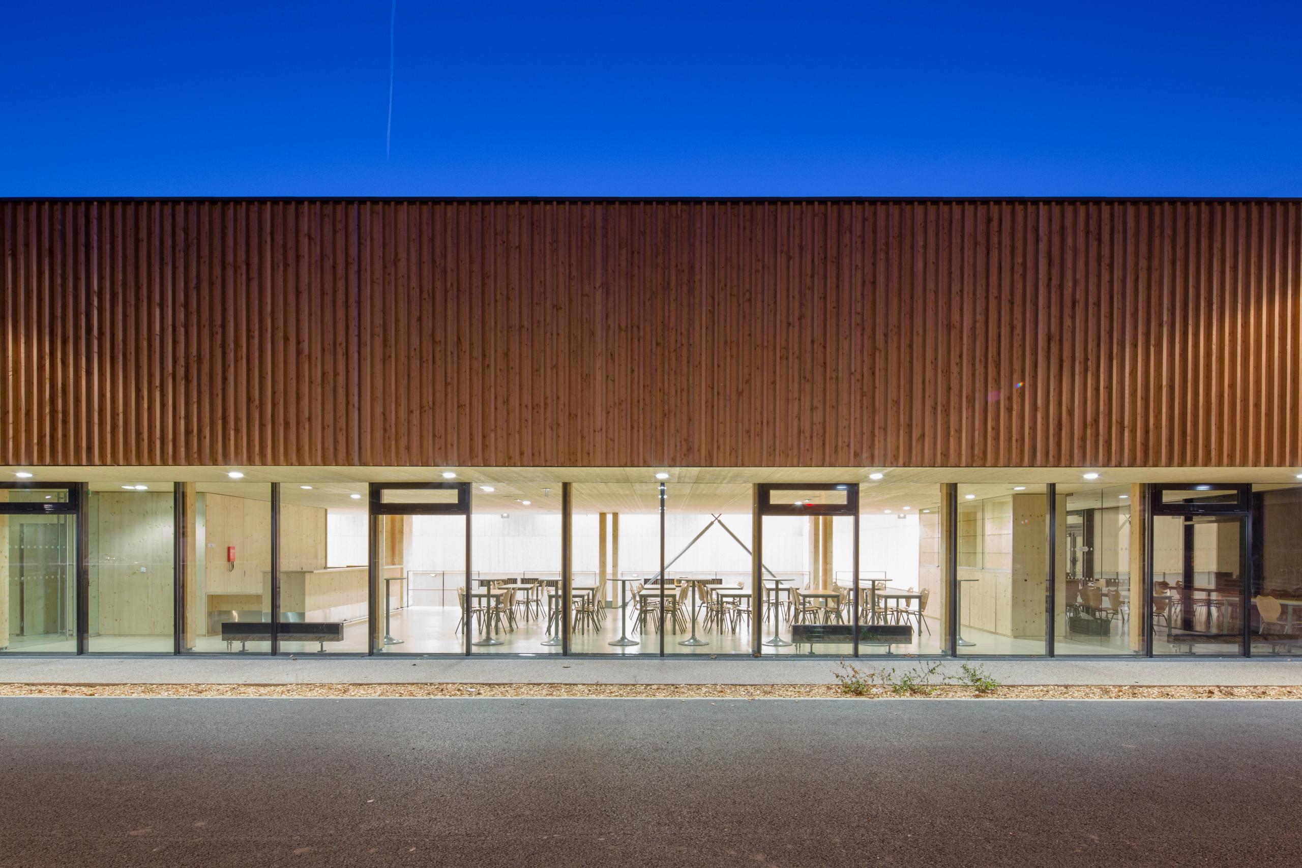 Façade nocturne du gymnase Hacine Cherifi à Rillieux-la-Pape, volume en bois sur socle béton, grande paroi vitrée révélant l’espace intérieur traversant.