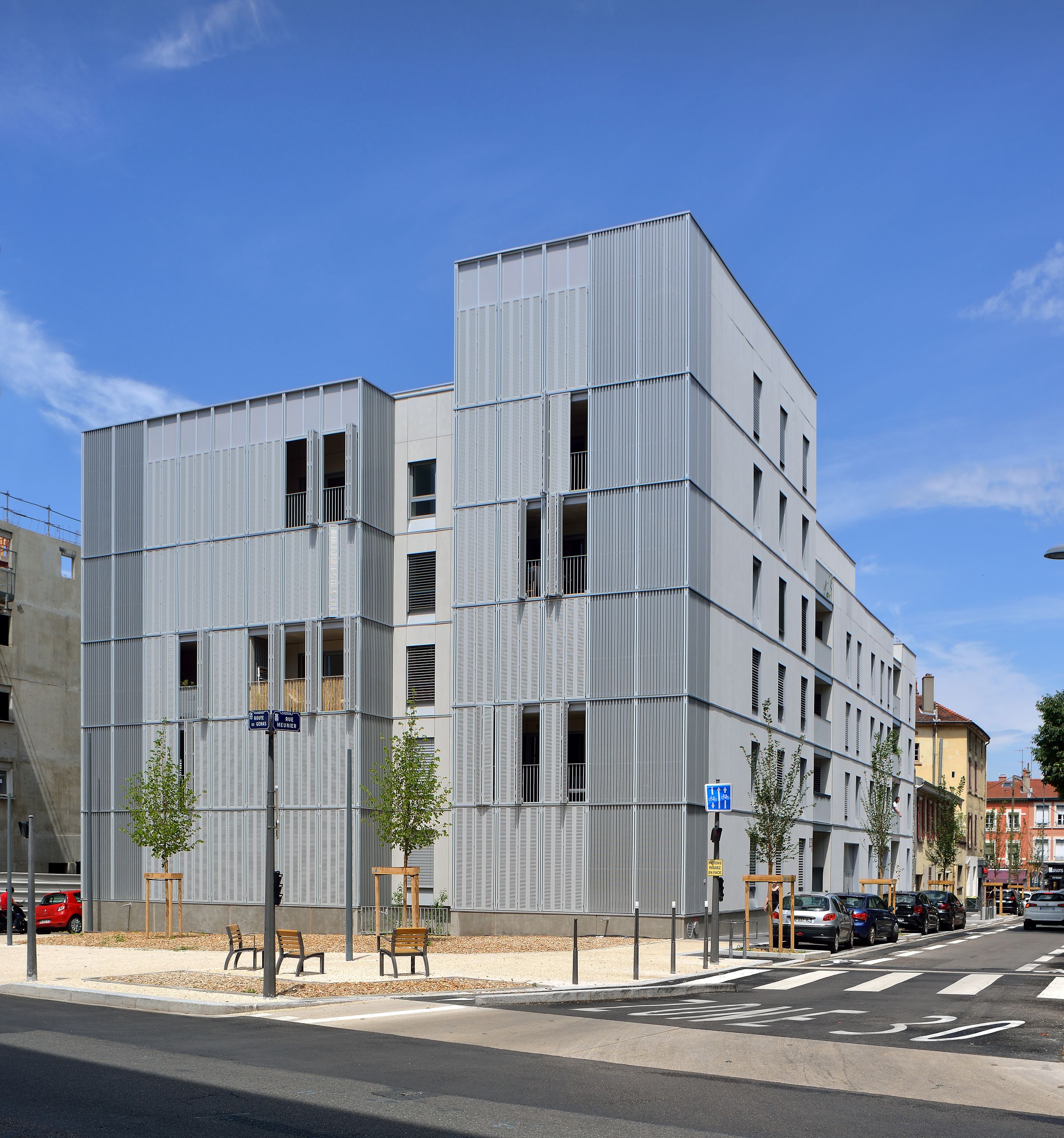 Vue de la résidence de 28 logements à Villeurbanne, volumes dégressifs en bardage métallique perforé, loggias en retrait et jardin surélevé sur rue.