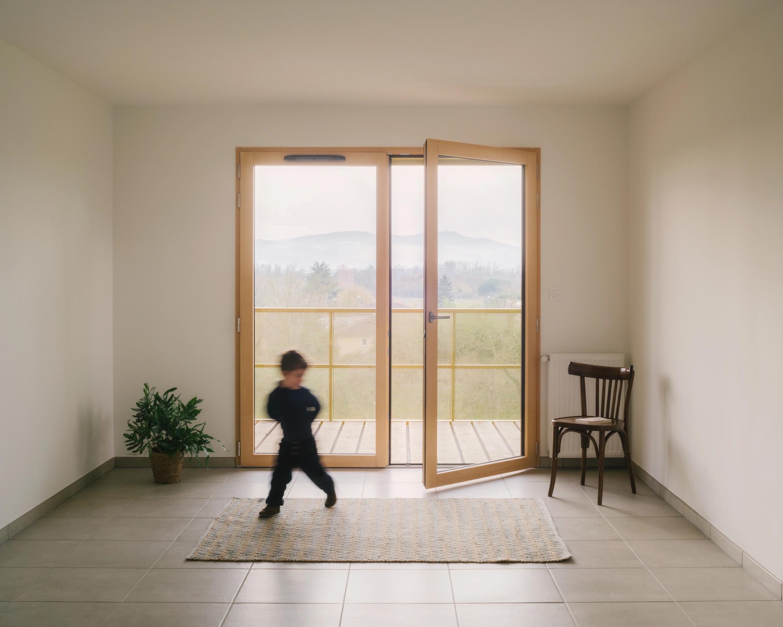 Intérieur d’un logement des 84 logements à Trévoux, grande baie vitrée bois ouvrant sur balcon, sol carrelé, paroi blanche et aménagement minimal.