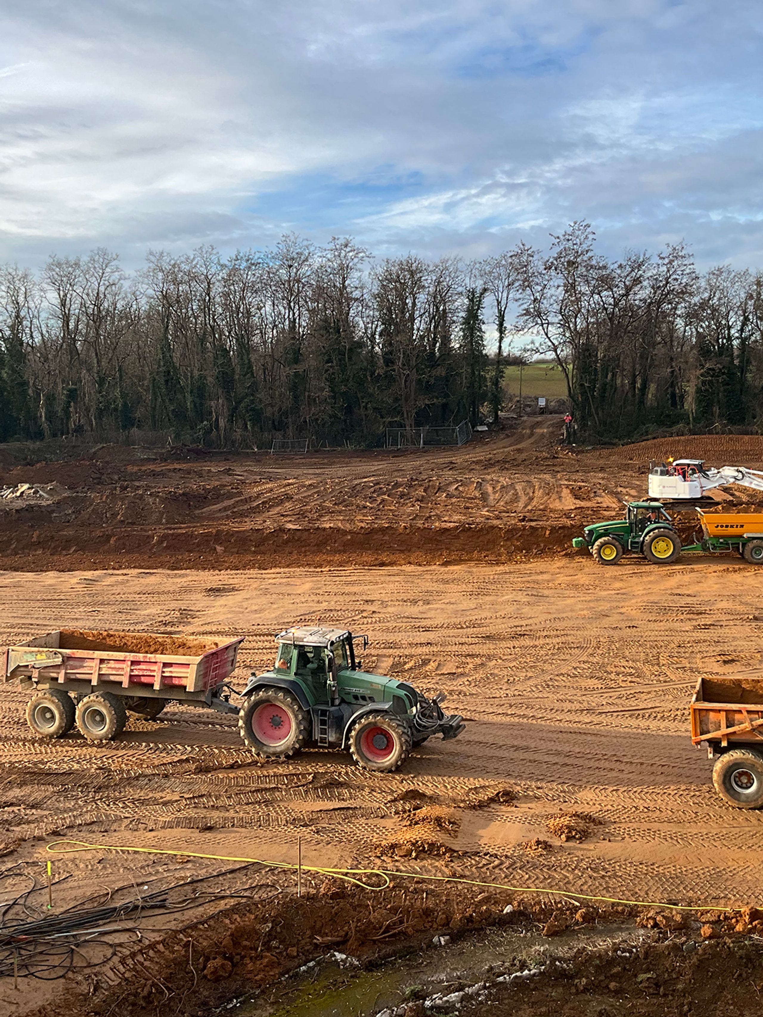 Earthworks for the Auxerre nursing home by Tectoniques, with tractors grading a large cut platform at the edge of a wooded greenway.