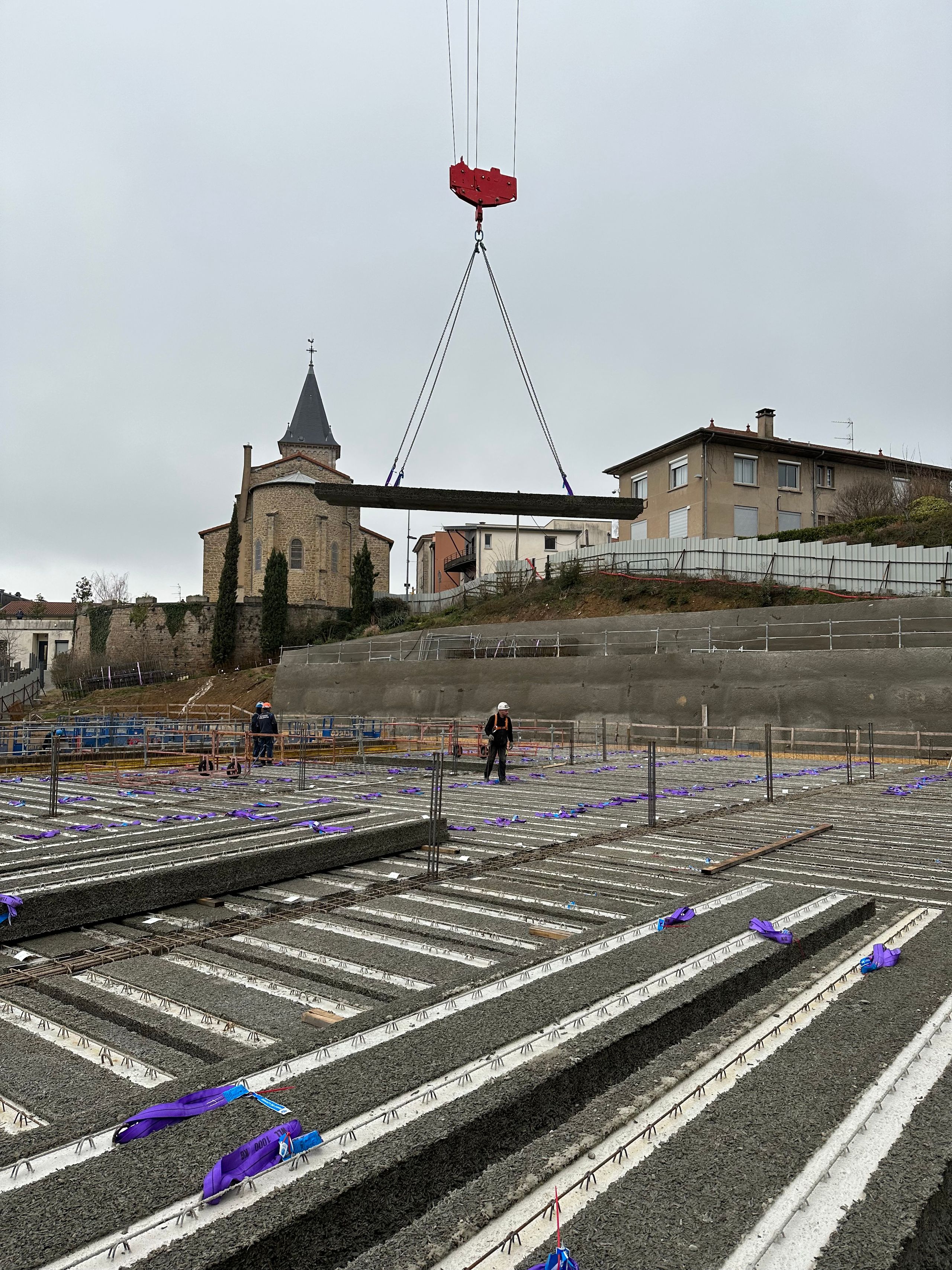 Chantier de la Maison des familles à Limonest : pose par grue d’une poutre béton sur planchers en béton de bois en terrasses, au pied de l’église du bourg.