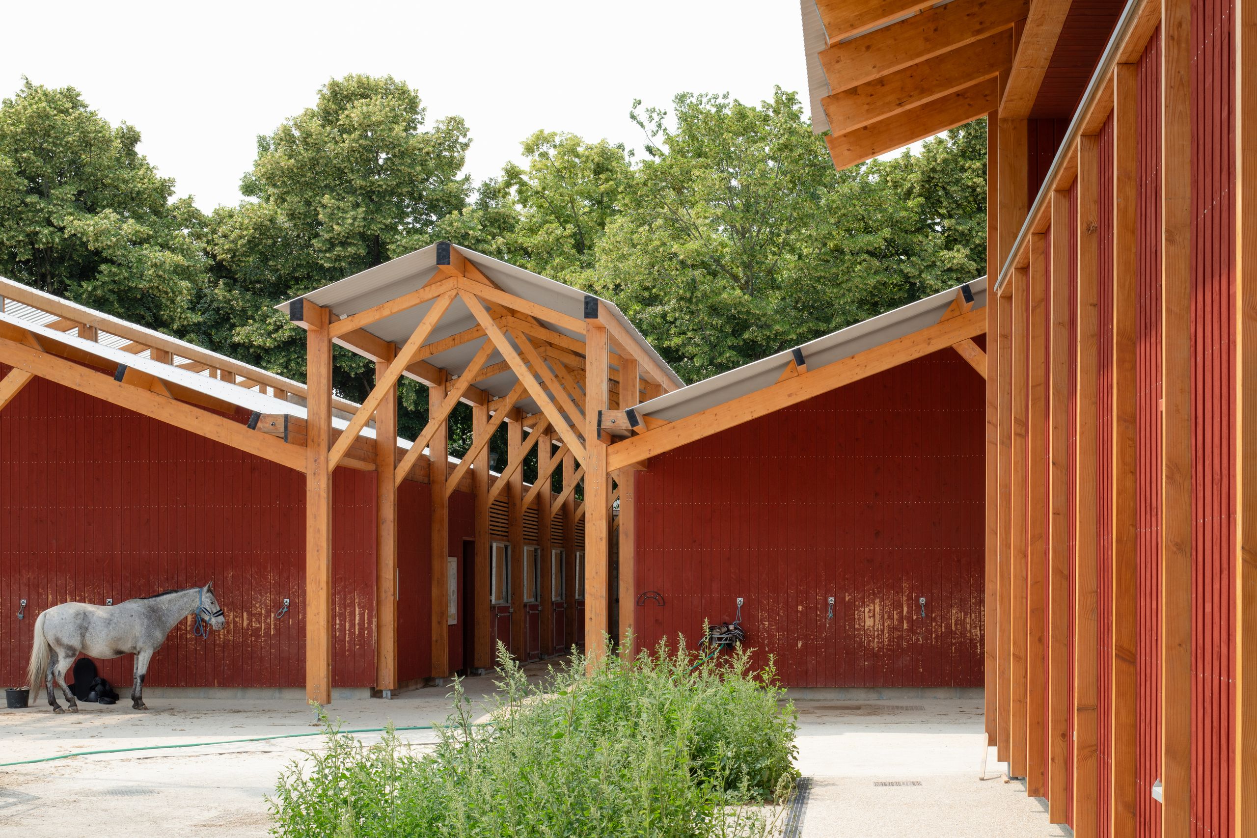 Centre équestre de Gennevilliers, bâtiments en bois bardés de rouge de Falun, charpente apparente formant porche couvert entre écuries et cour plantée.