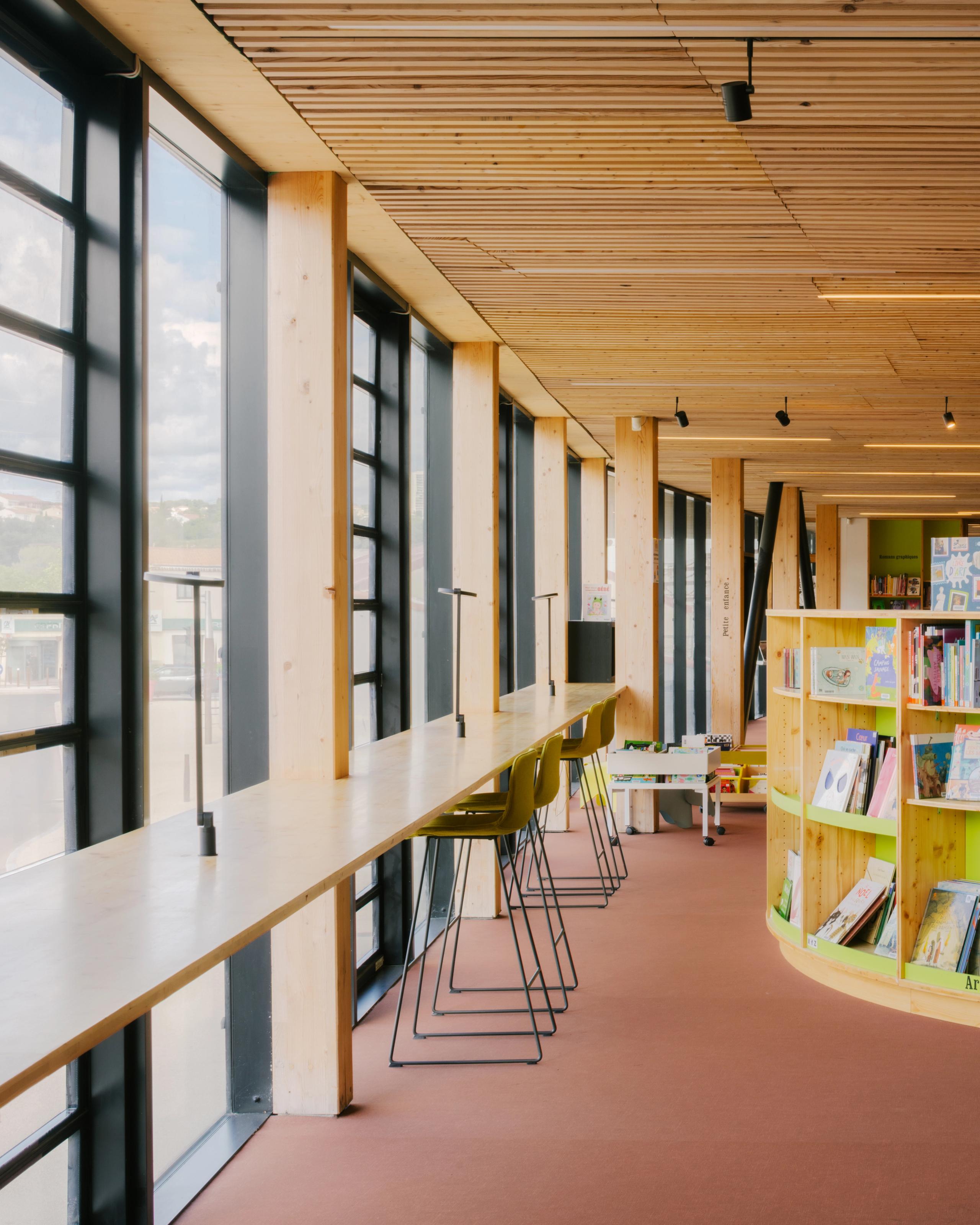 Intérieur de la médiathèque du groupe scolaire Claudie Haigneré aux Pennes-Mirabeau, espace de lecture en bois lumineux avec façade vitrée et mobilier intégré.