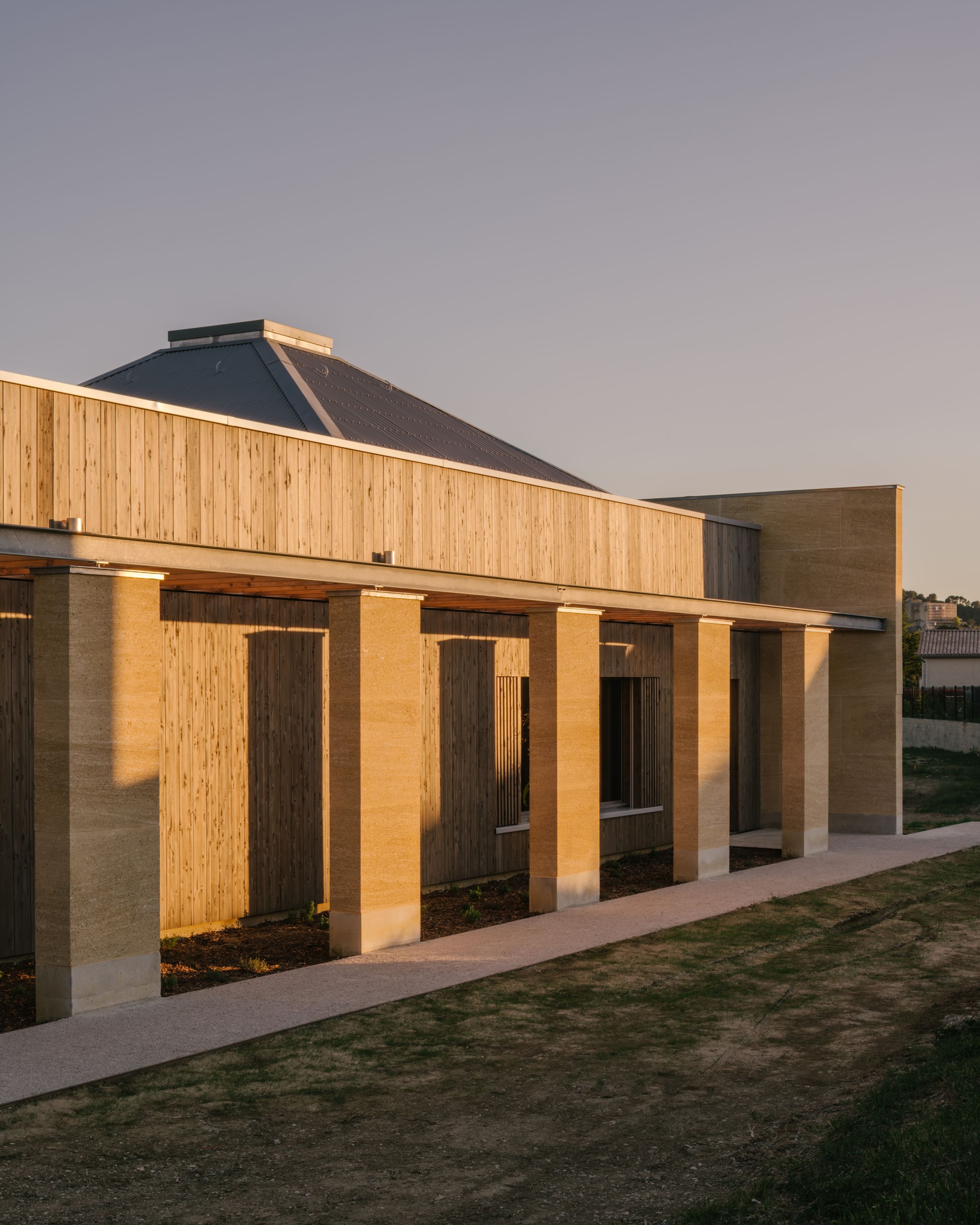 Façade du pôle culture et jeunesse de Vaison-la-Romaine : portique en pierre massive, bardage bois vertical et volume de toiture émergent au-dessus de l’enceinte.