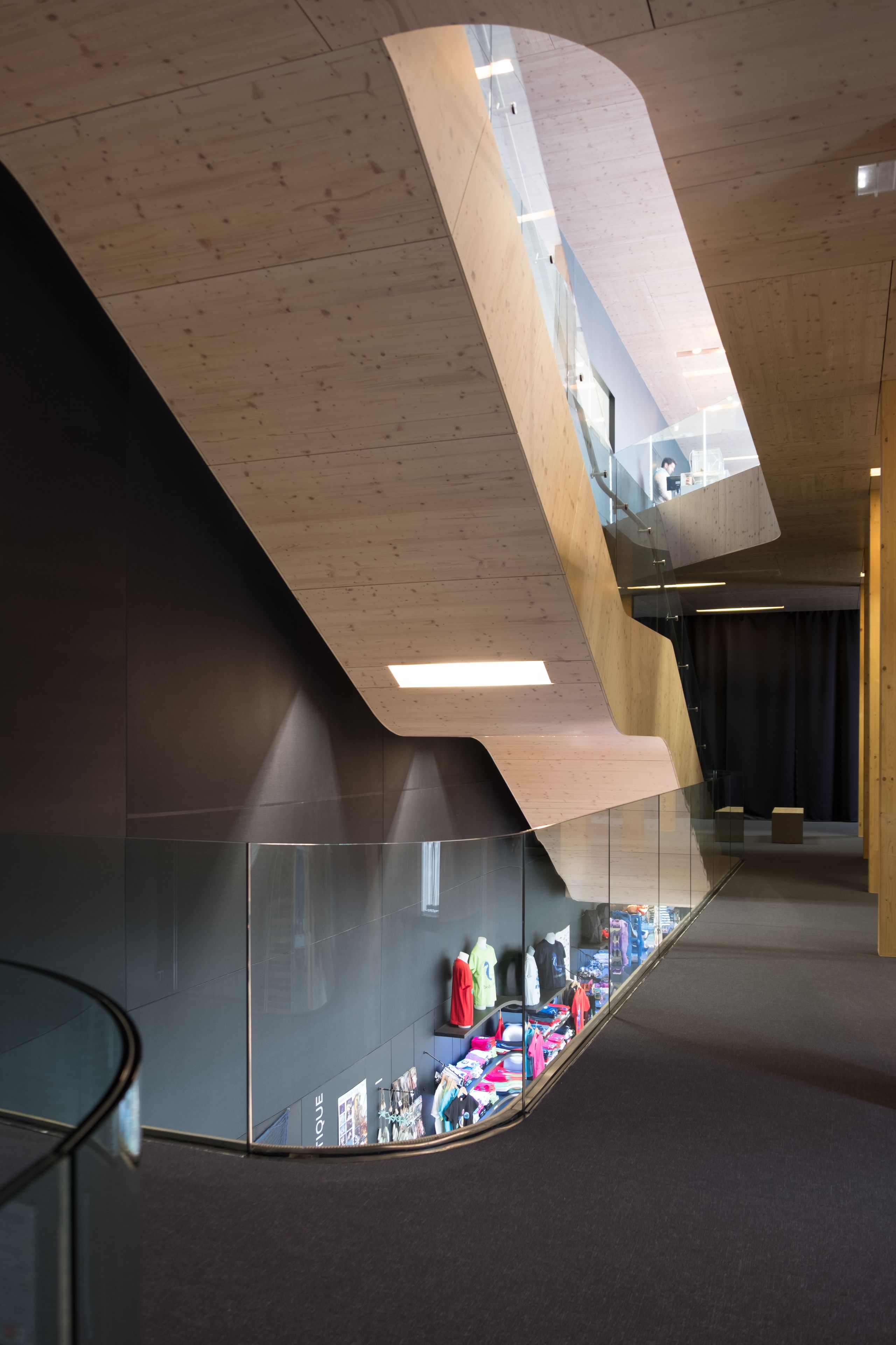 Intérieur du Biodiversarium à Banyuls-sur-Mer : circulation verticale sculptée en bois lamellé, garde-corps vitrés et parois sombres surplombant l’espace d’exposition.