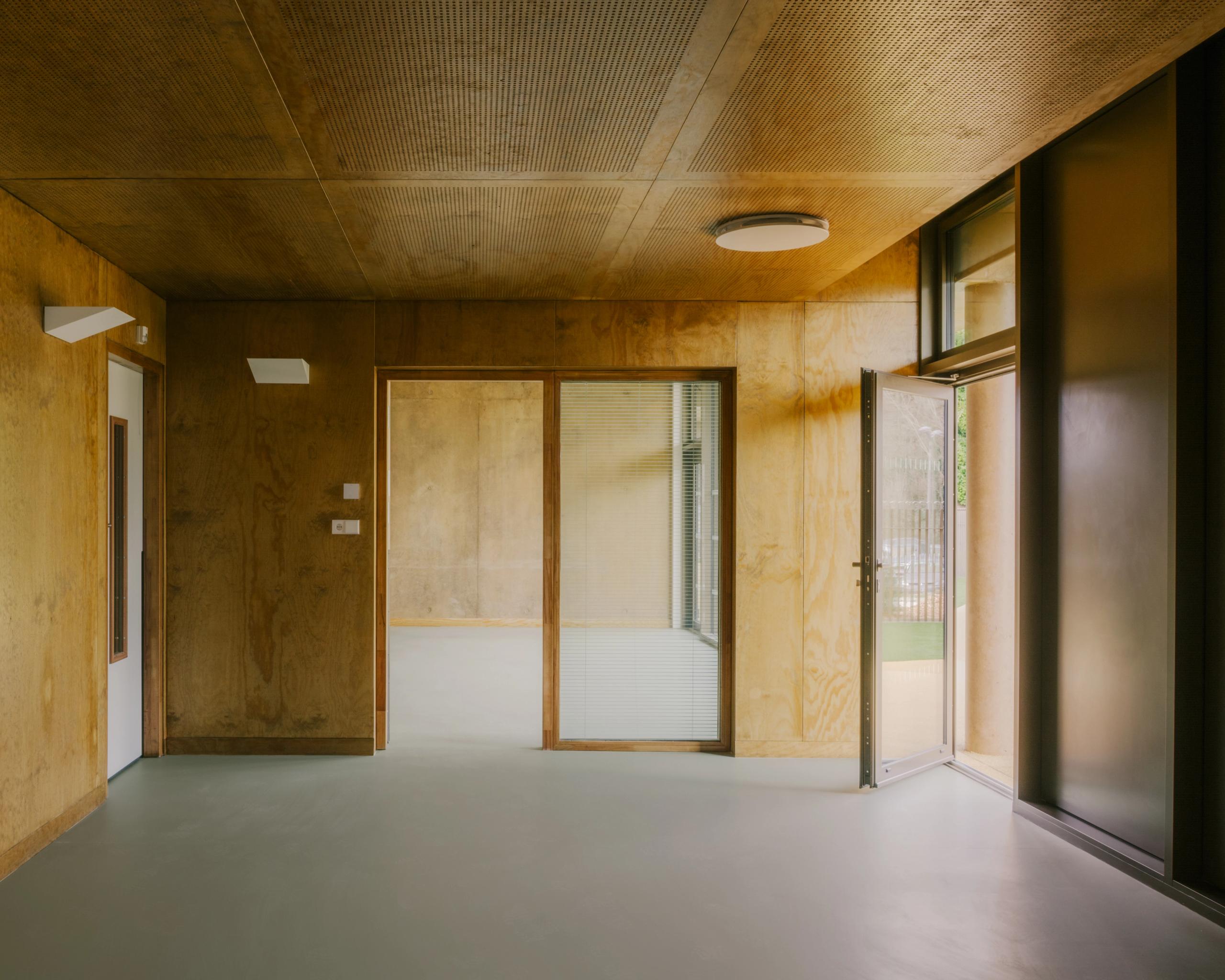 Intérieur de la Maison des familles à Limonest : plateau en plan libre, parois et plafond en panneaux bois, sol lisse clair, large baie vitrée ouverte sur l’extérieur.